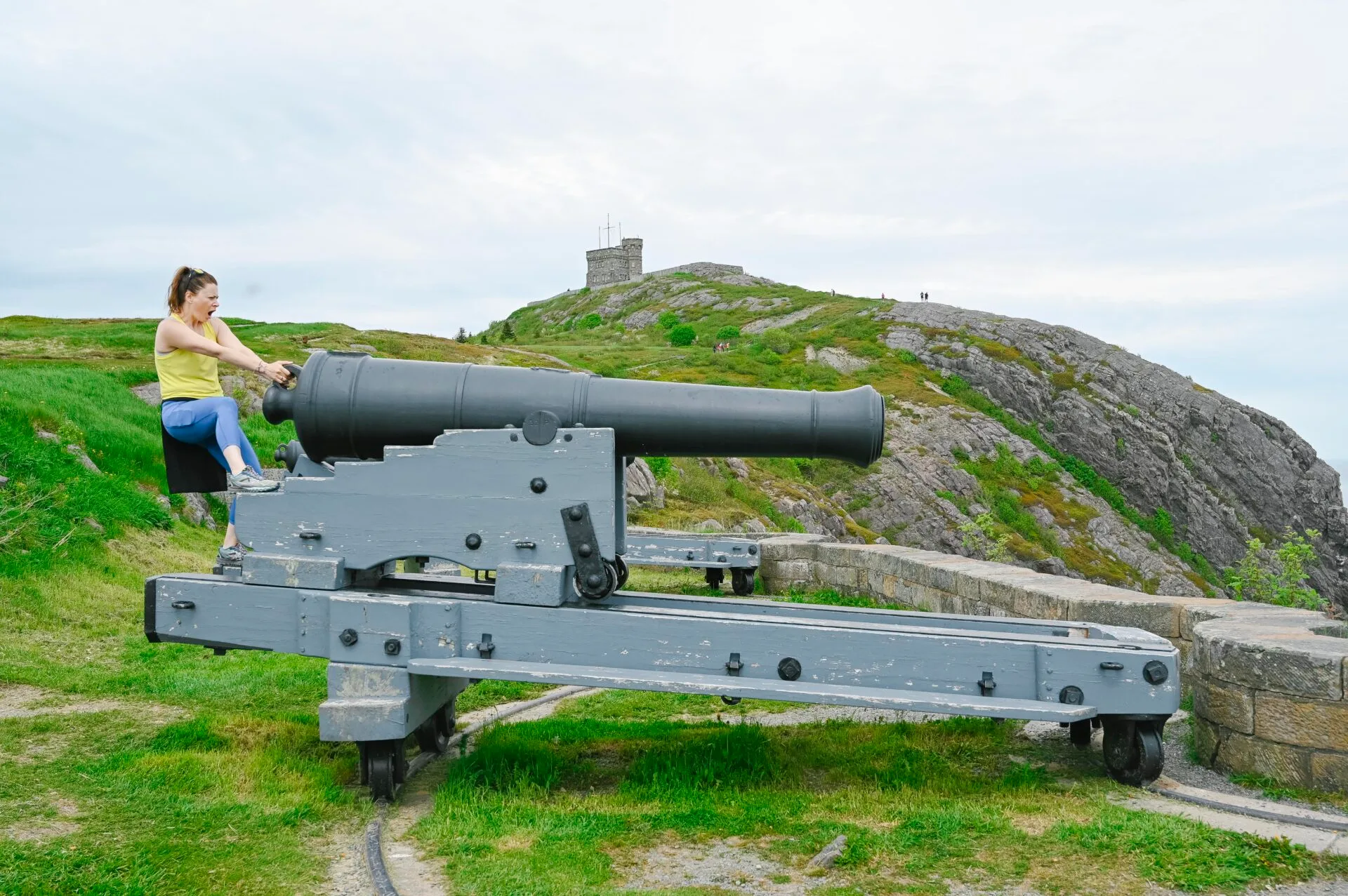 a woman wearing a yellow shirt sits at the firing end of a large cannon at signal hill national historic site