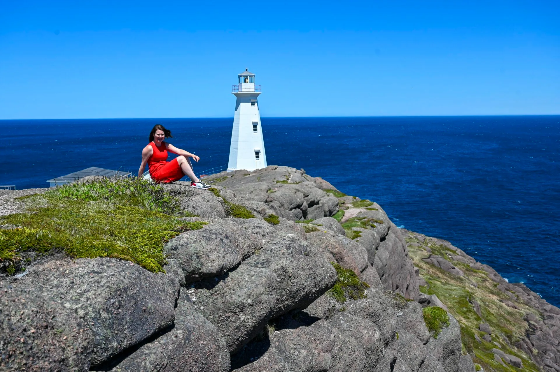 a woman in a red outfit sits on the rocks in front of the cape spear lighthouse