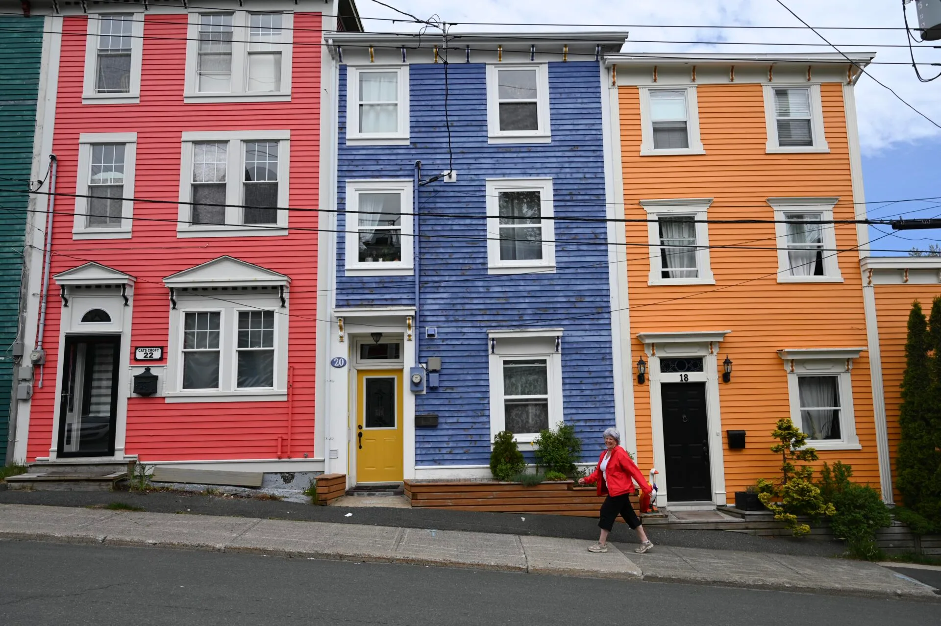 a woman wearing a bright red coat walks past the jellybean row houses, painted a pink, blue and orange, in st.john's