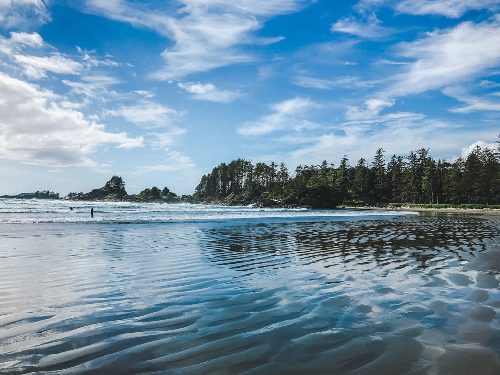 view of someone standing in the ocean waves under the sun and blue skies on cox bay beach in tofino