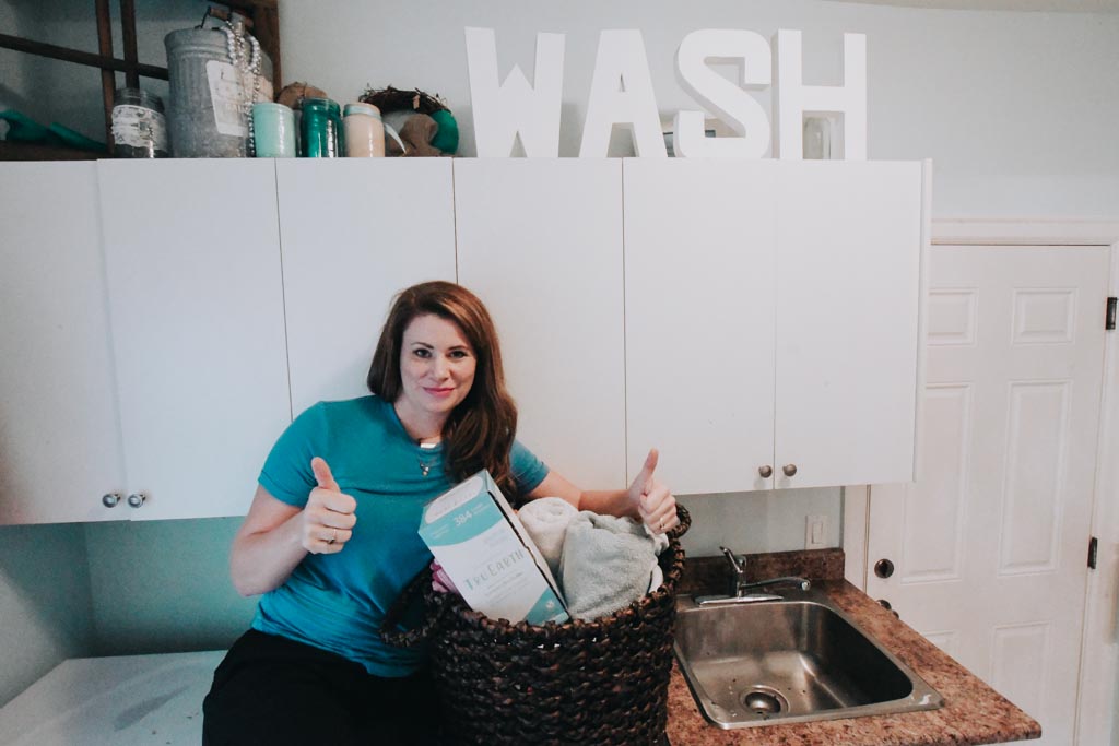 woman giving two thumbs up as she sits on her laundry machines with a basket of laundry and tru earth laundry strips beside her