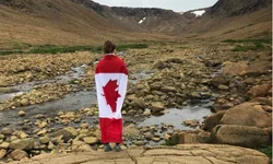 woman standing with a canada flag on her back looking out towards a creek and mountains