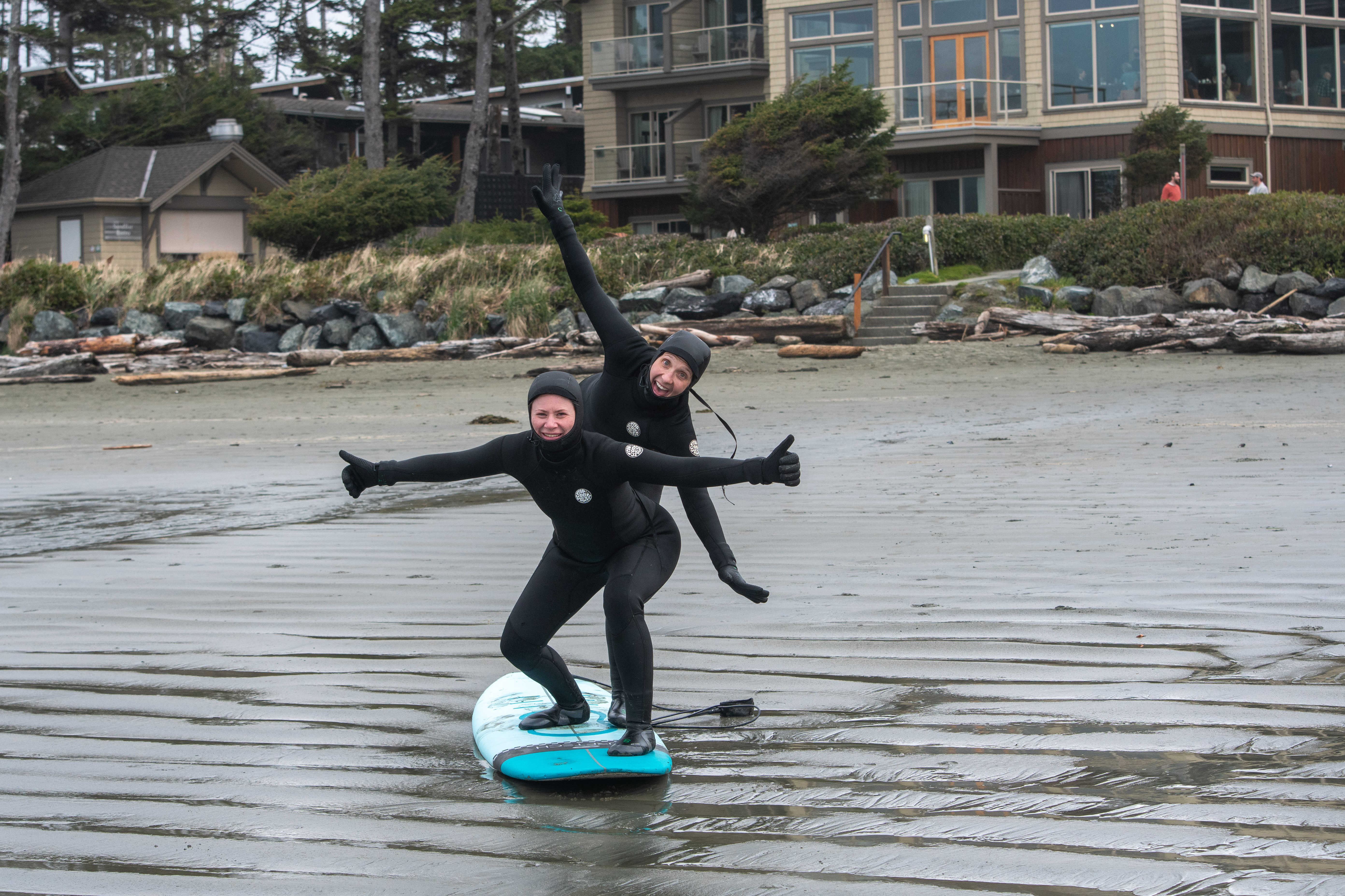 2 women wearing wetsuits and standing on a surf board on the beach acting silly
