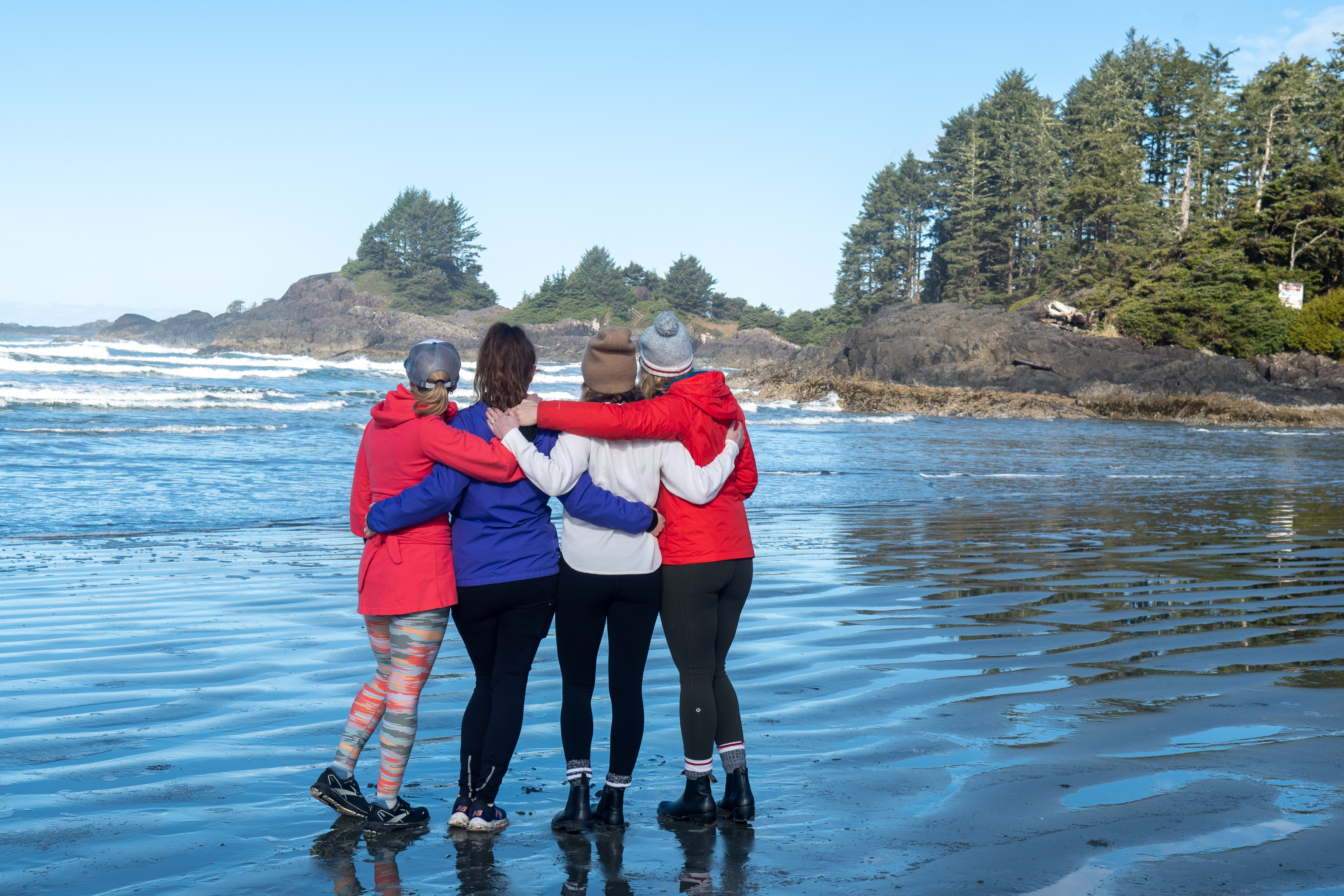 4 women facing away from the camera with their arms around each other and looking out at the ocean on a beach in tofino