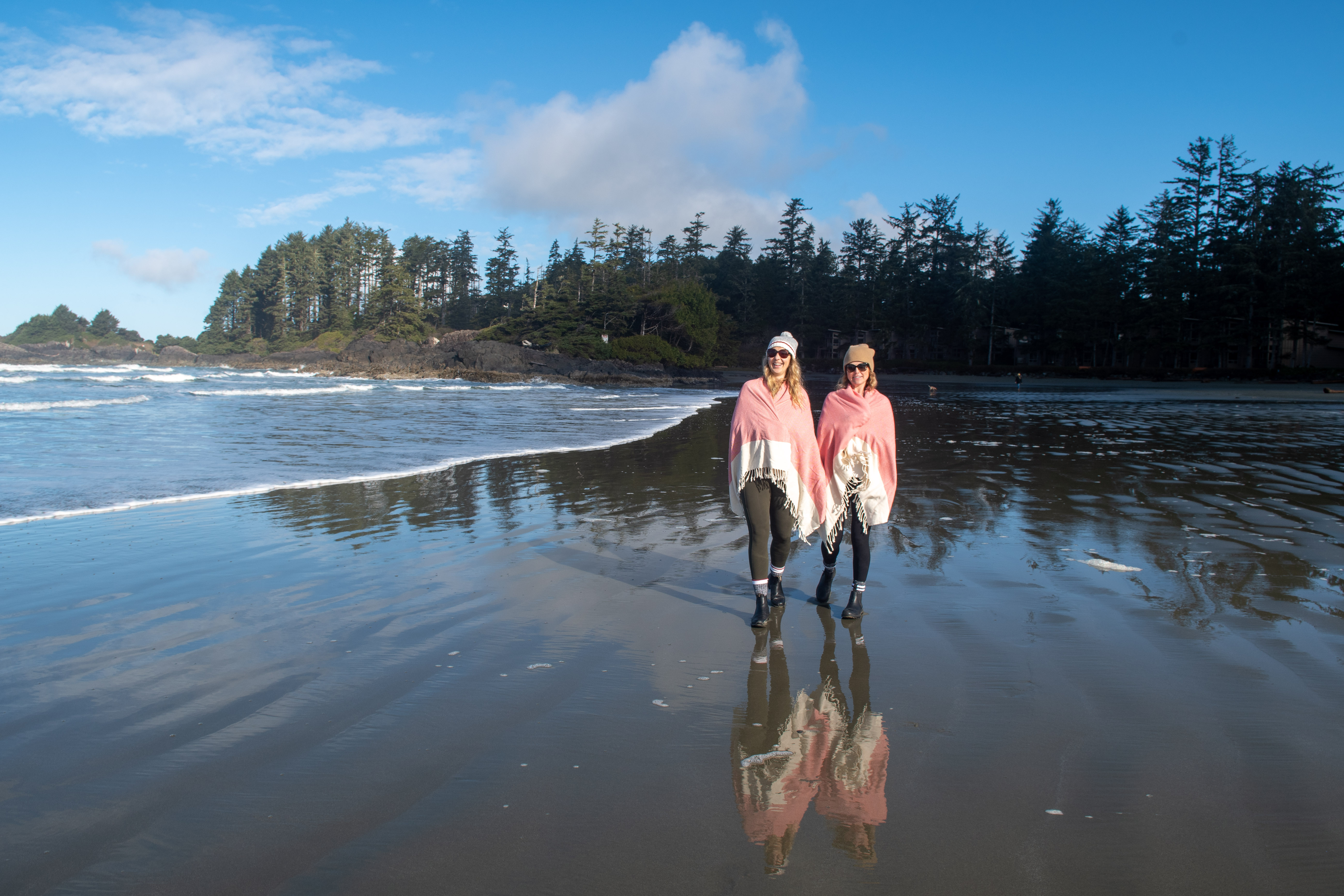 two women with blankets draped on their shoulders, stand on a beach in tofino