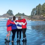 4 women facing away from the camera with their arms around each other and looking out at the ocean on a beach in tofino