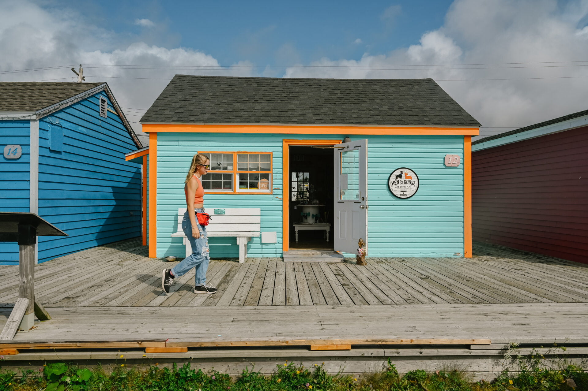 woman walking along the boardwalk at fisherman's cove nova scotia past a blue building with orange trim with a sign that reads hen and good pet supply co.