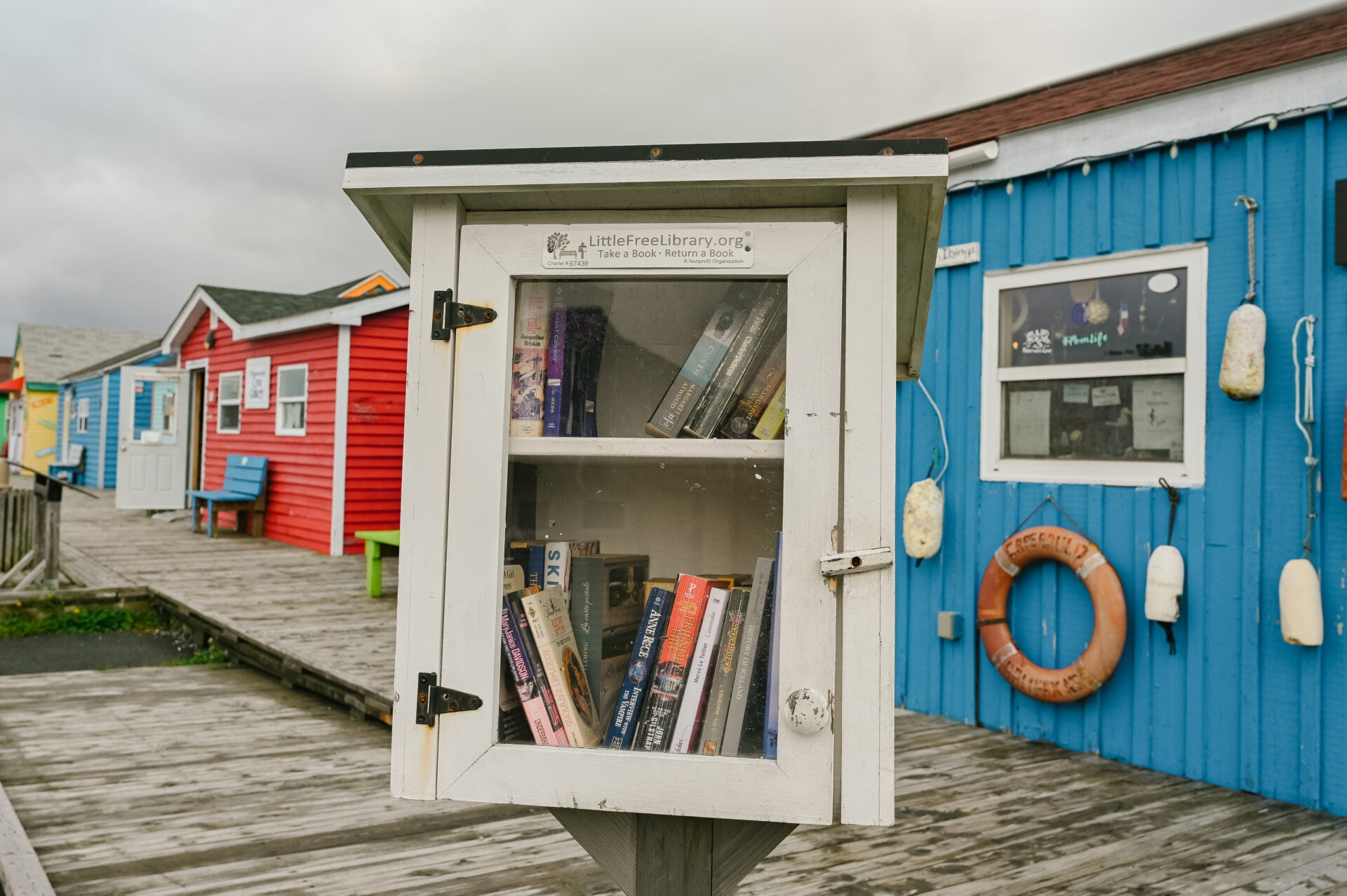 close up of a little free library stand with a row of shops along the fisherman's cove nova scotia boardwalk behind it