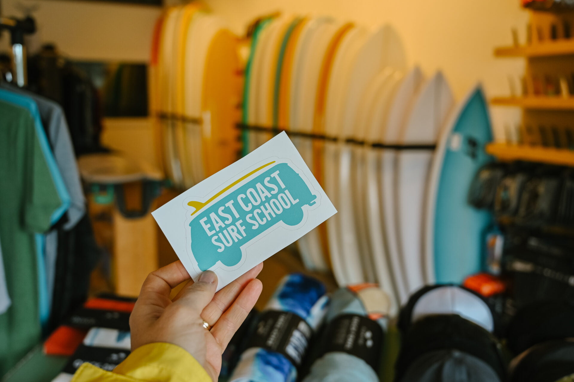 hand holding a east coast surf school sticker, surf boards in the background