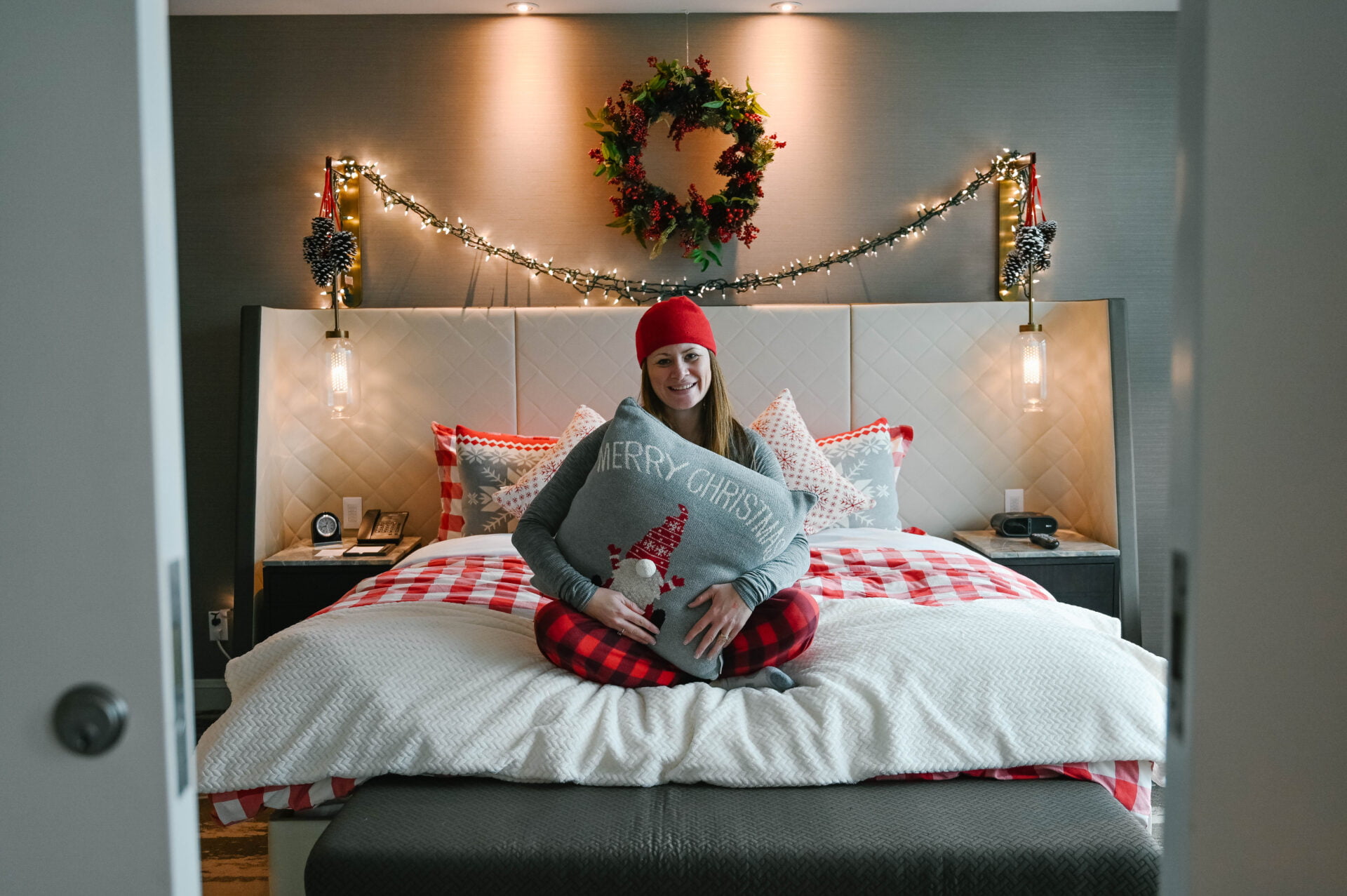 woman sitting crossed legged on a bed holding a merry christmas pillow with a gnome of it.