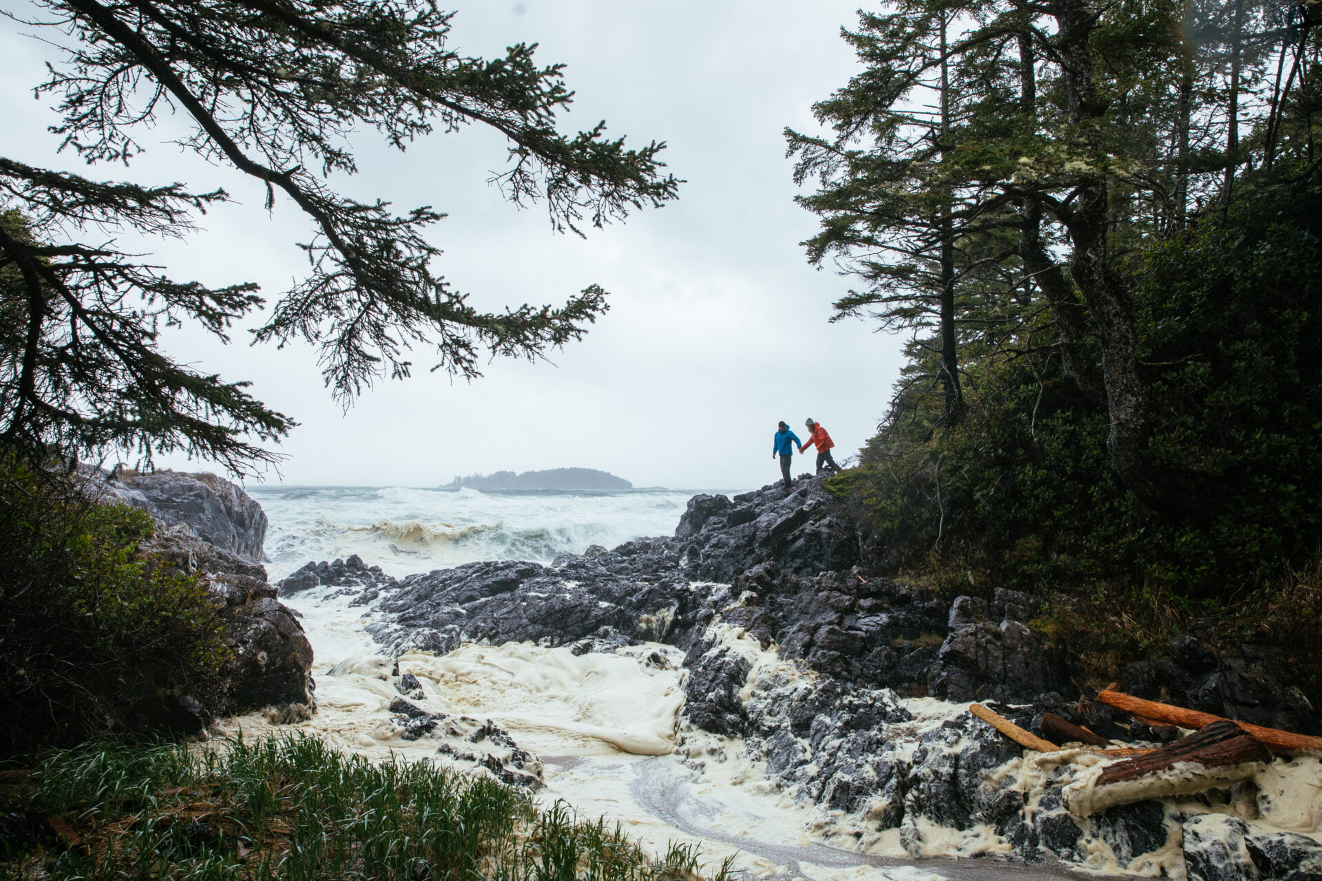 a couple holds hand while climbing on the rocks beside a very stormy ocean at chestermans beach in tofino on vancouver island