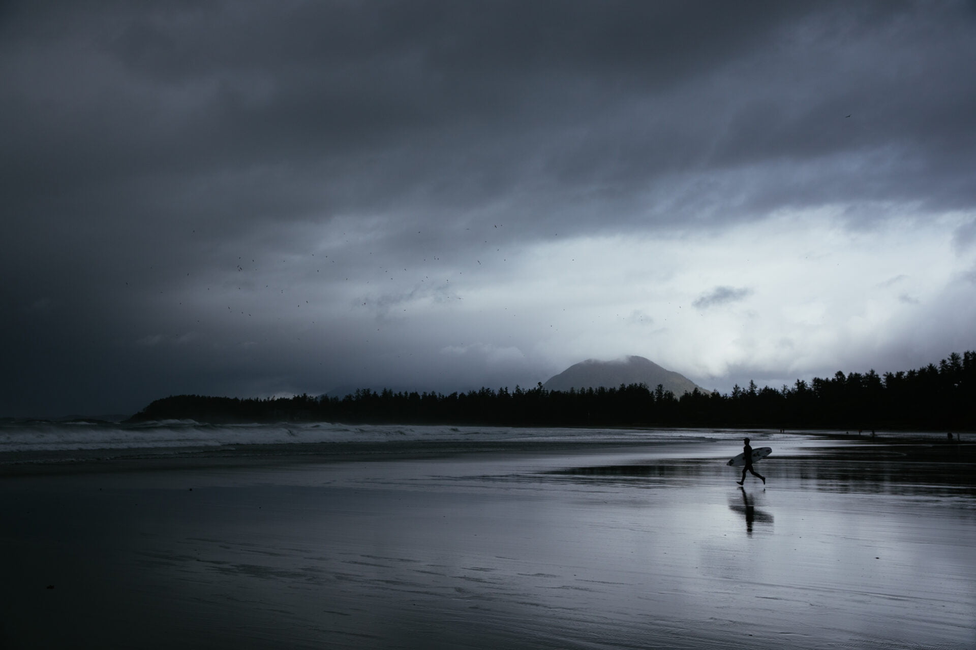surfer carrying their surf board across chesterman beach, heading out towards the waves, on a dark and stormy day