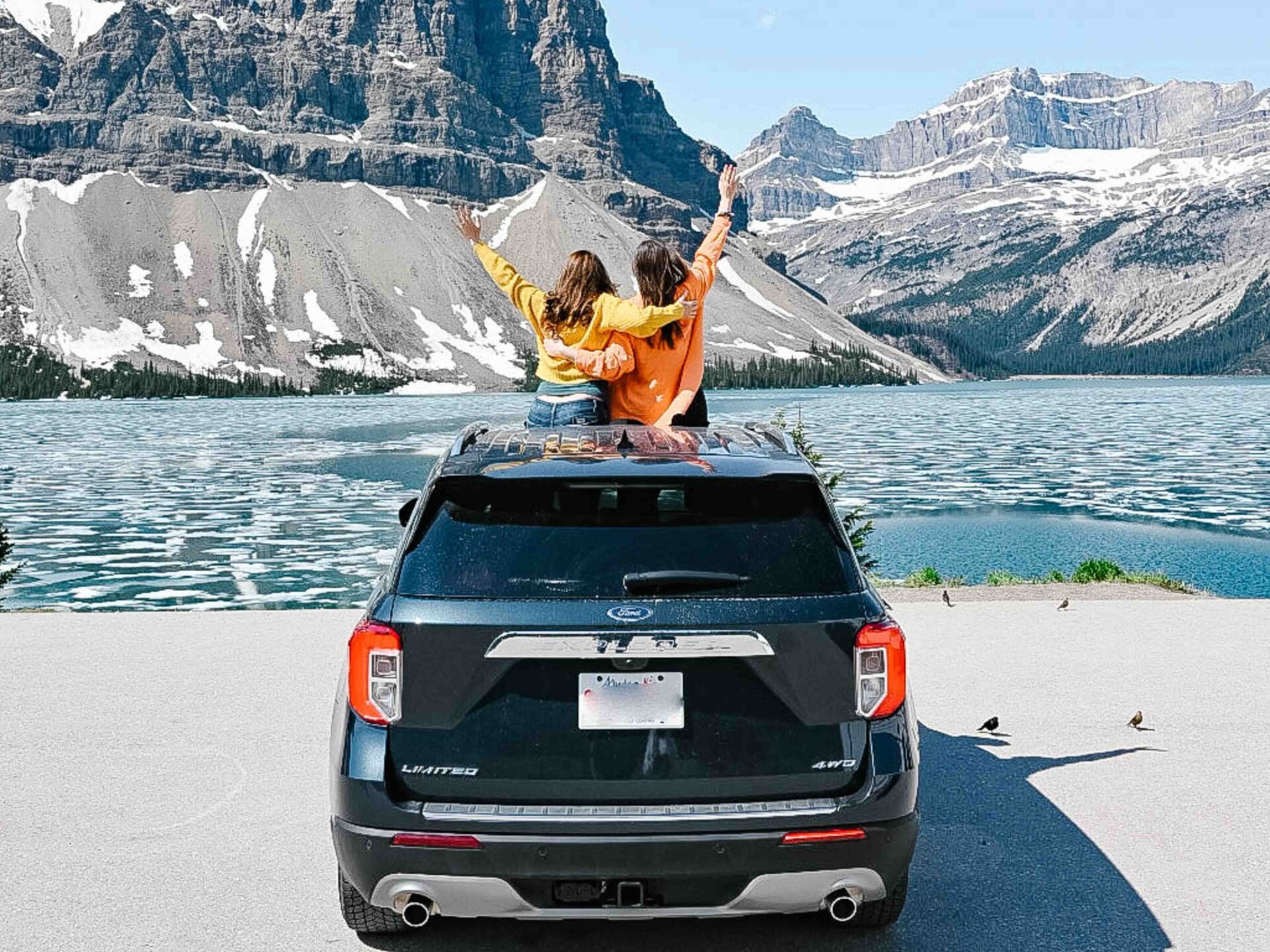 two woman standing out the top of the sunroof of a black SUV with bow lake in the background on their banff to golden road trip