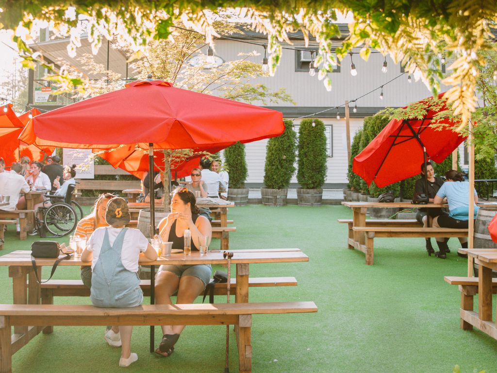 lots of people enjoying food and drinks while sitting on picnic tables under red umbrellas at trading post brewing in fort langley, one of the best patios langley