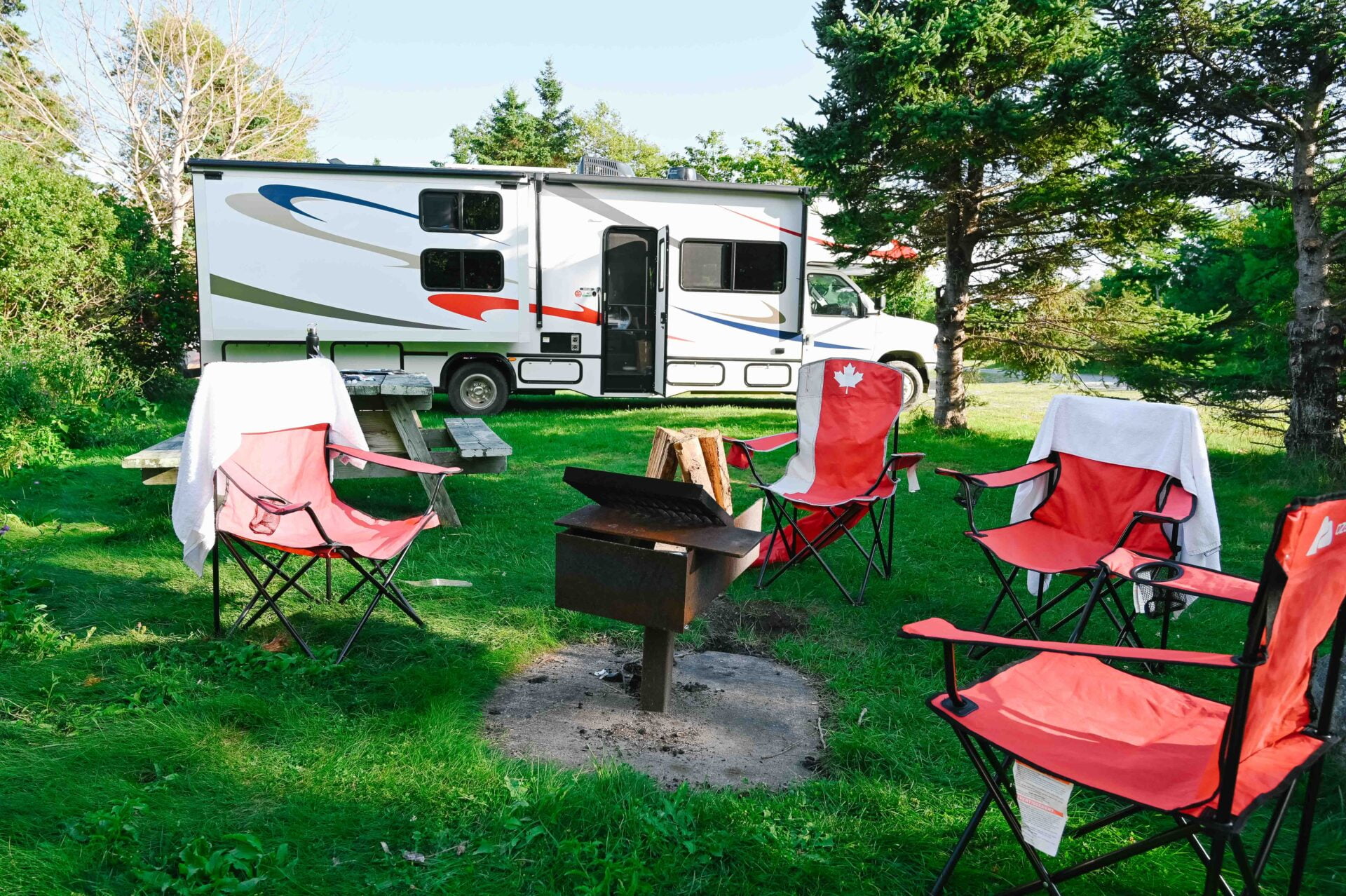 an RV sits in the background of a campsite where camping chairs are placed around the campfire