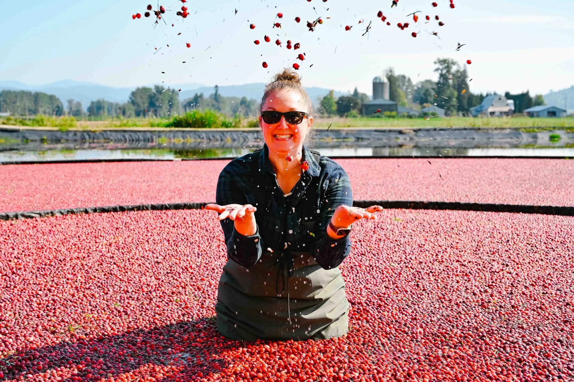woman standing in the cranberry field throwing cranberries in the air, she has a big smile and is wearing a plaid shirt