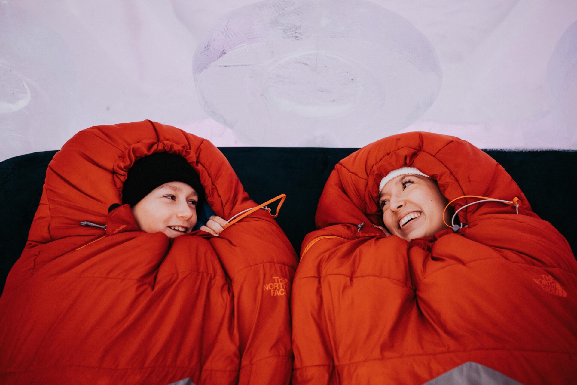 a couple in mummy style winter sleeping bags look at each other while lying on a bed inside the Hôtel de Glace