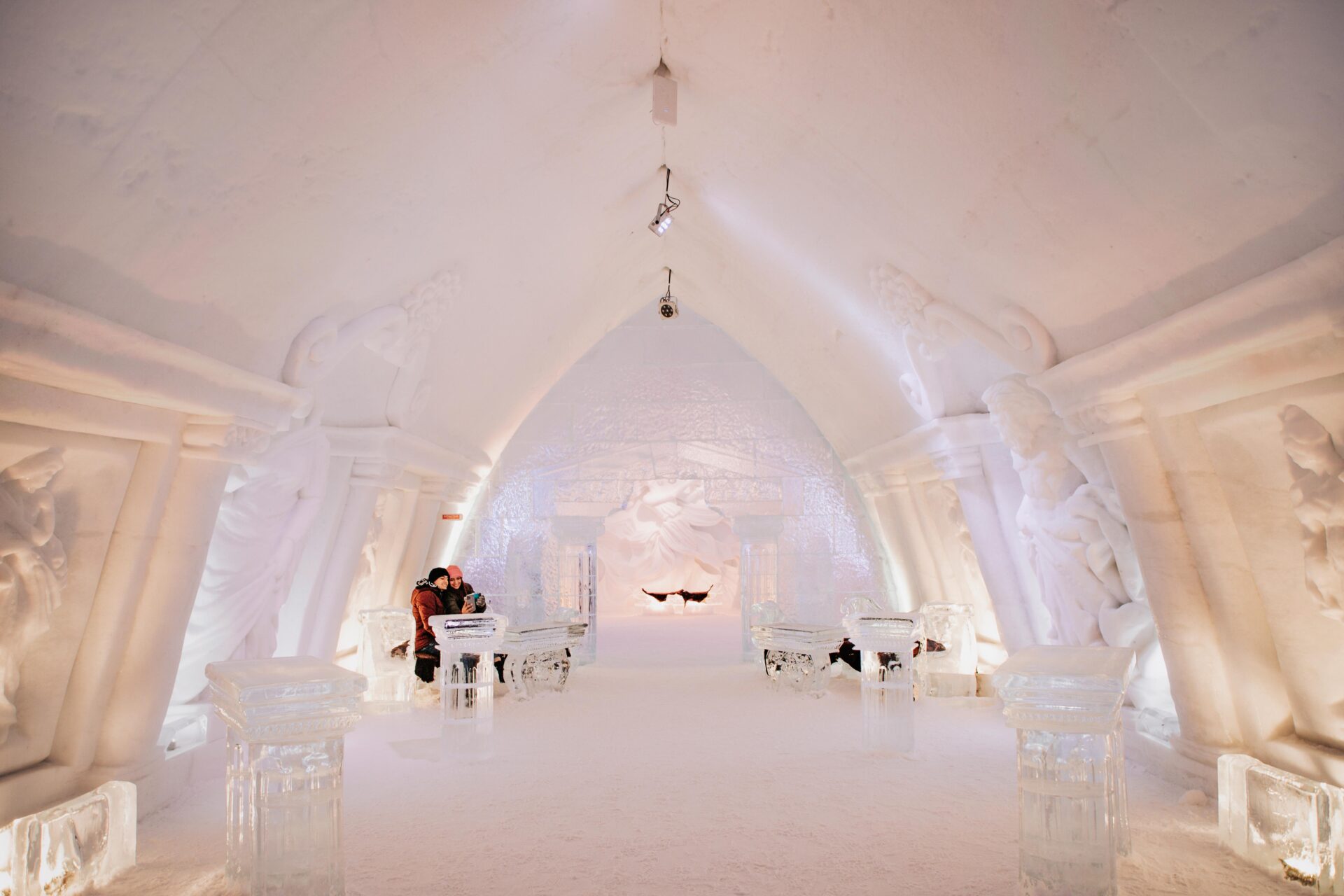 a couple sit on an ice bench while taking a selfie inside the Hôtel de Glace