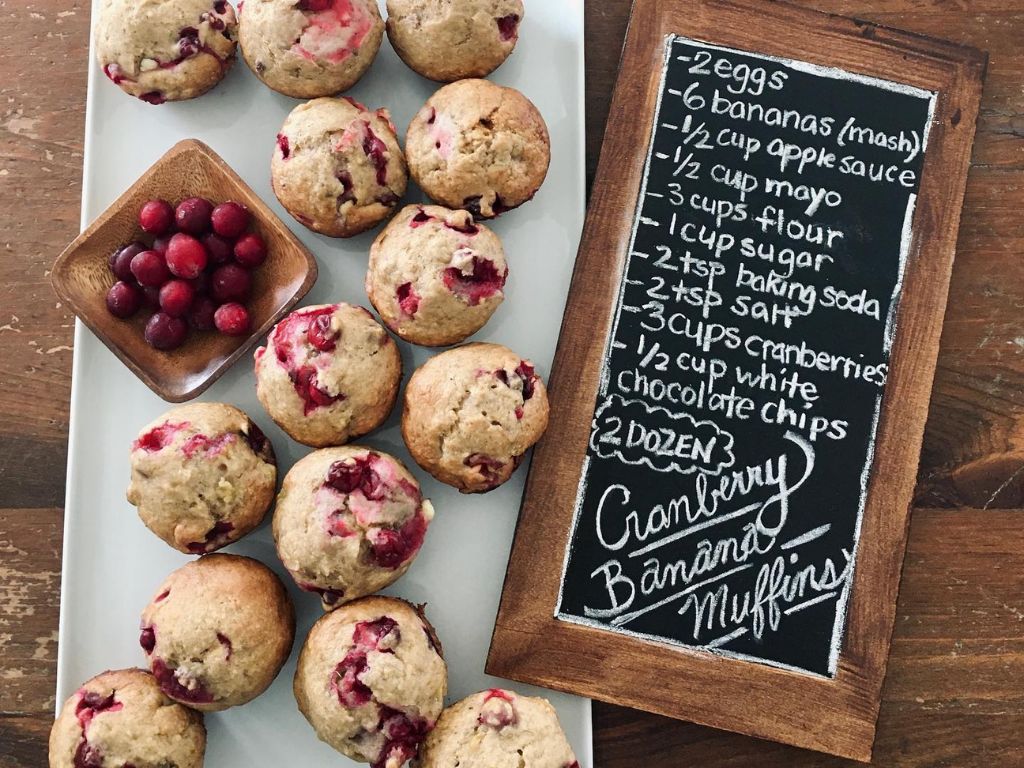 cranberry muffins on a plate, with fresh cranberries in a bowl, with the muffin recipe printed on a small chalkboard beside the plate