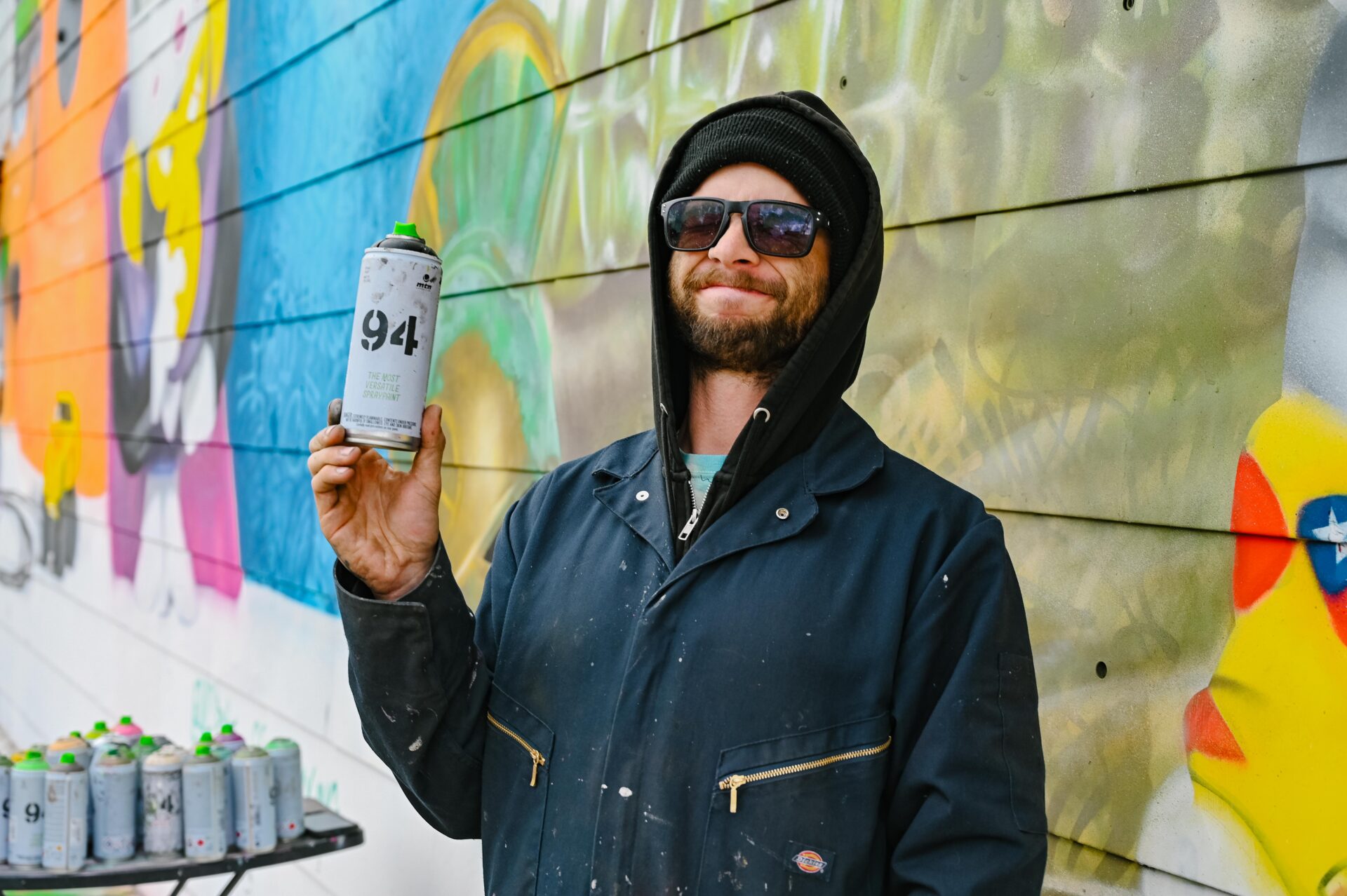 Artist Aaron - AG_PNT, holds a can of spray paint, in front of his large mural that is in progress