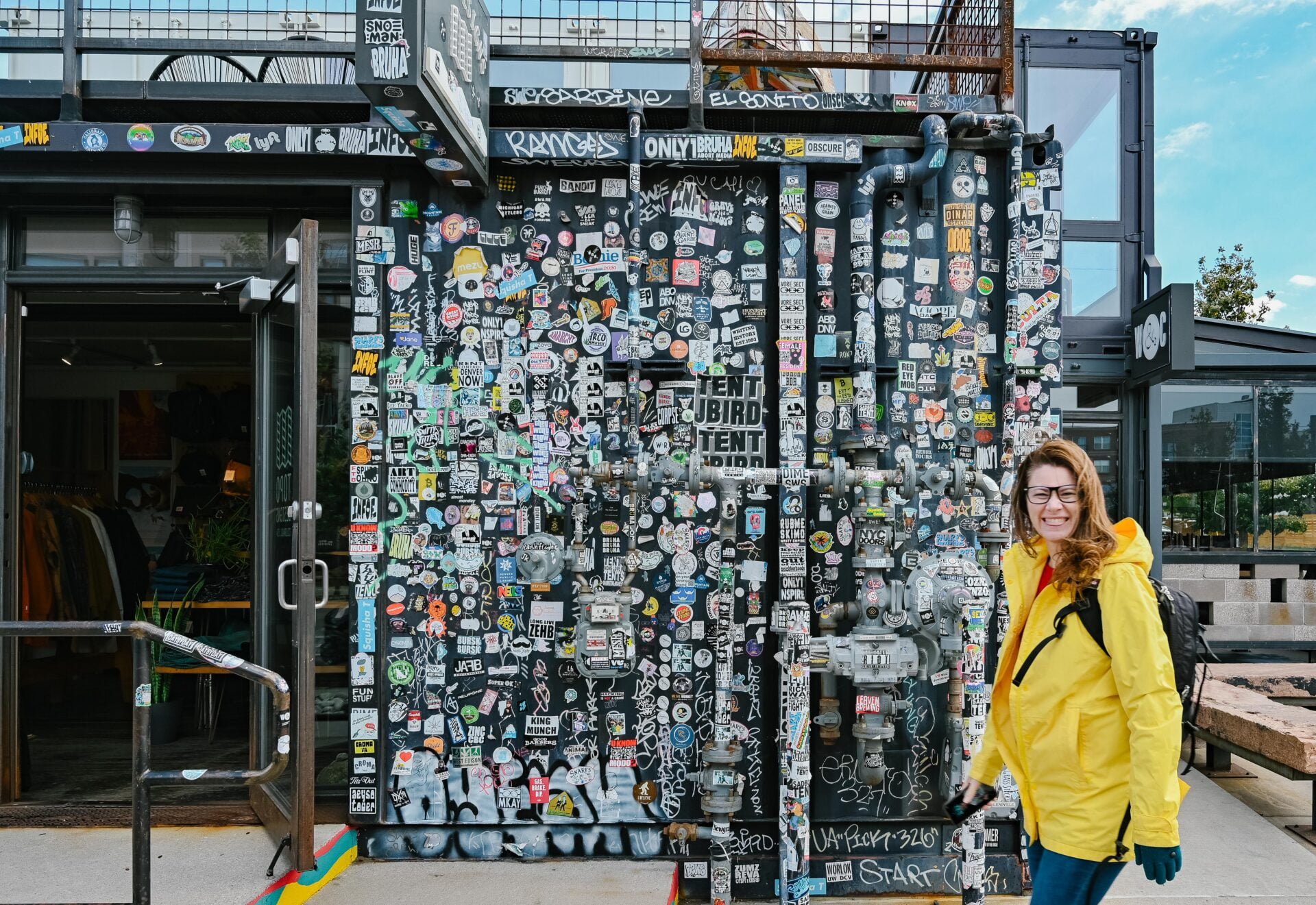 a woman in a yellow jacket standing in front of a wall covered in stickers, found on the denver graffiti tour