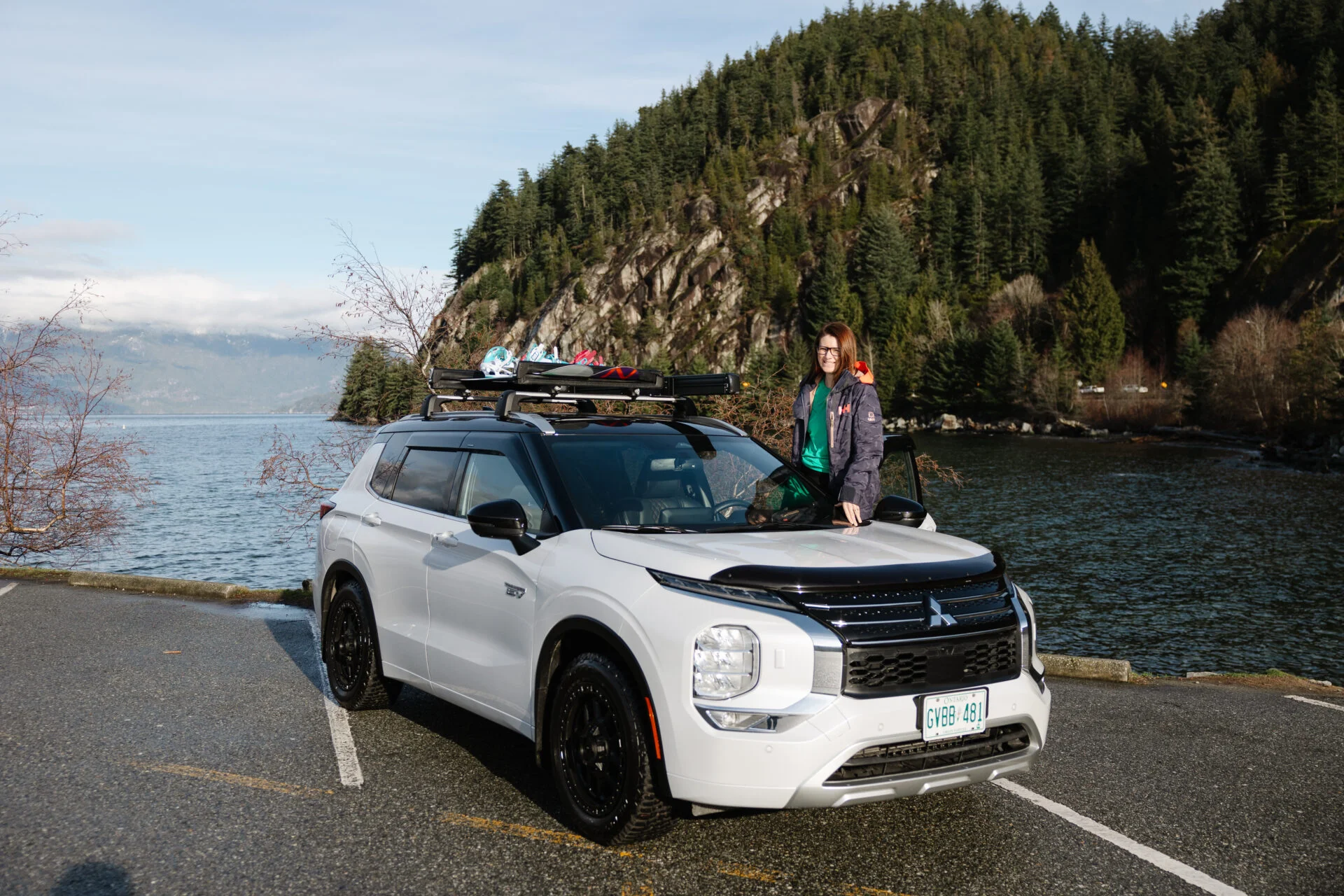 woman stands in the open door of a white mitsubishi outlander phev, mountains and the ocean are behind her