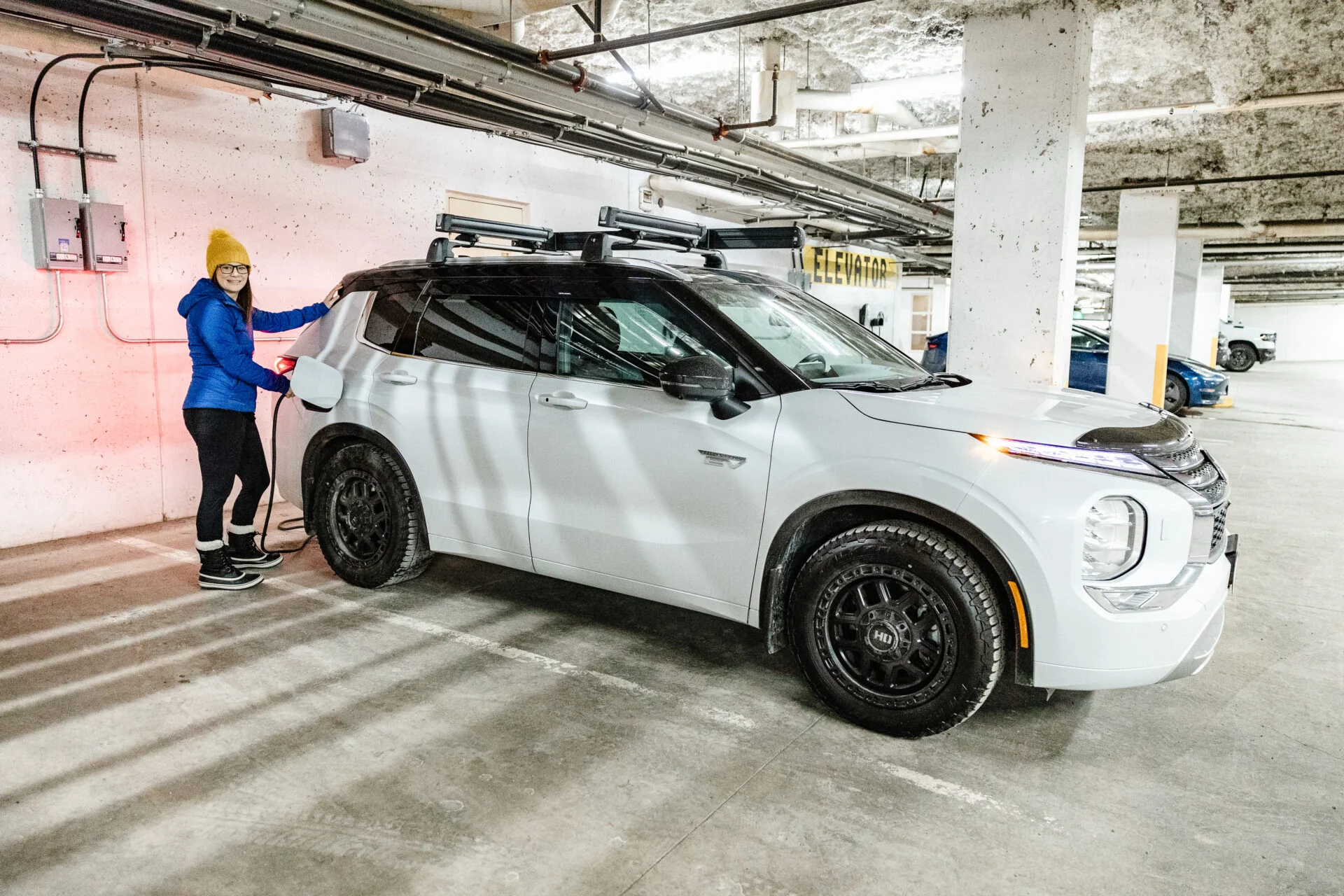 woman wearing a blue coat and yellow toque is plugging in a white electric SUV in an underground parking garage