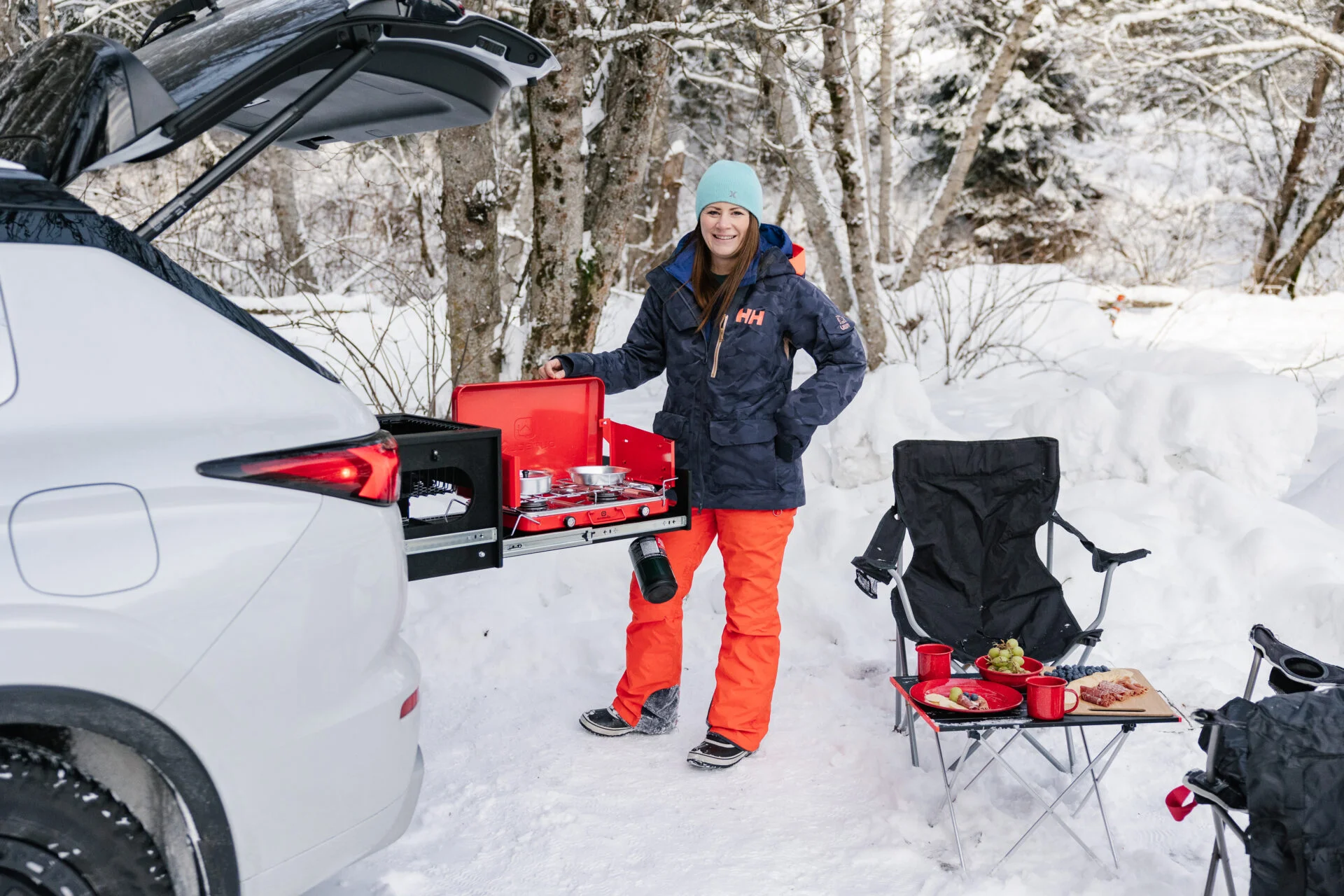 a woman dressed in warm snow gear stands at the back of a mitsubishi outlander phev, its nomad camp kitchen is slid out and she is cooking