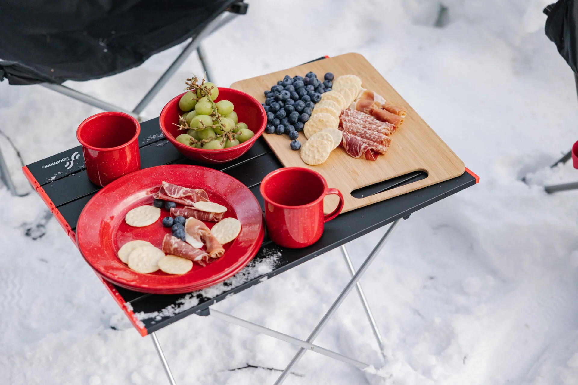 a small camp table sits in the snow, atop it is a charcuterie board of meat, cracker and fruit. a plate, bowl of grapes and mugs are also on the table.