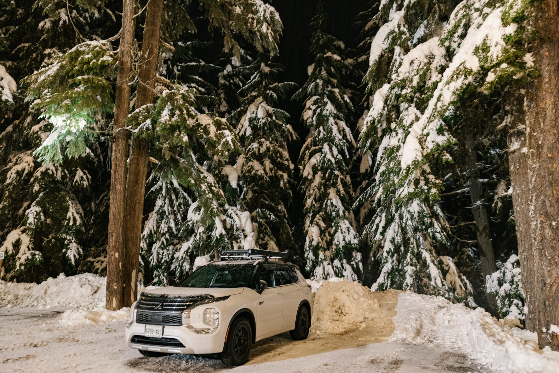 a mitsubishi outlander phev sits parked in front of a snow bank, large snow covered trees are behind it
