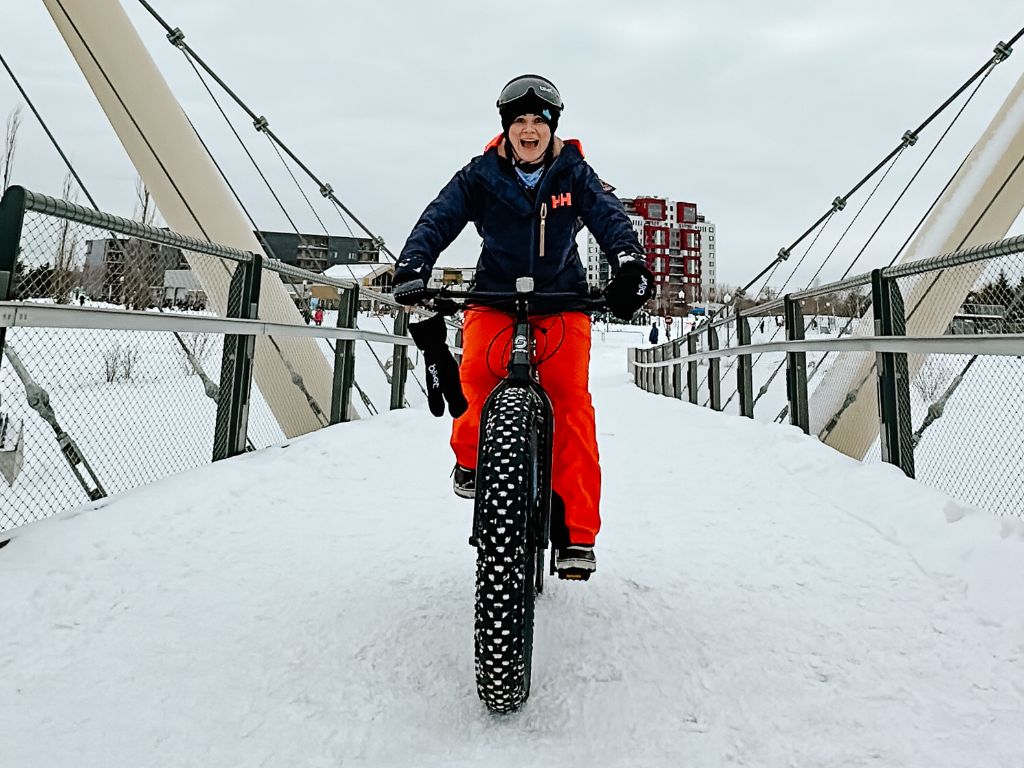 woman with huge smile on her face rides her Quebec city bike rental towards the camera over a bridge in the snow