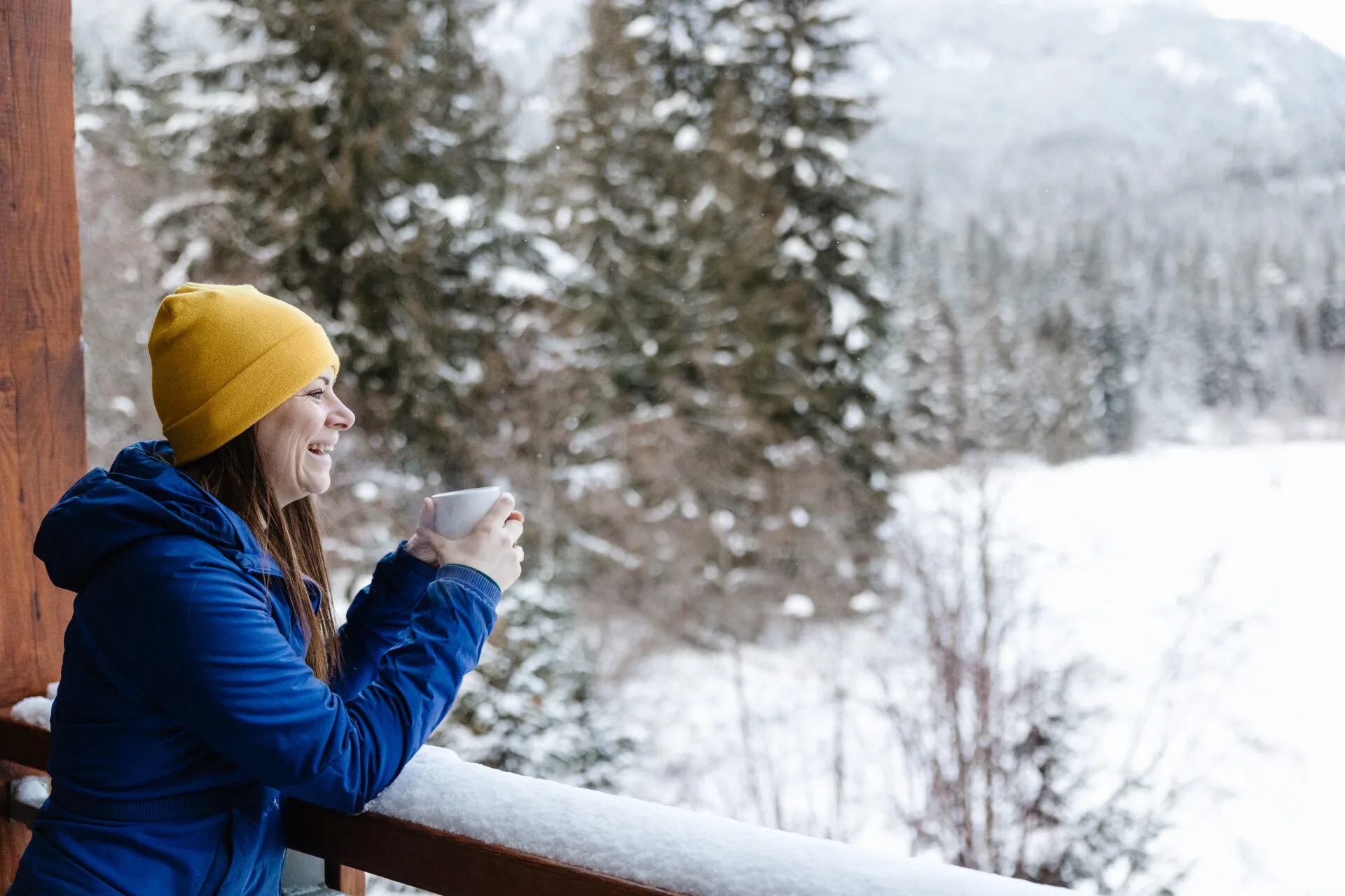 a woman wearing a blue coat and yellow toque drinks a coffee while overlooking the view from a hotel room at the nita lake lodge in whistler