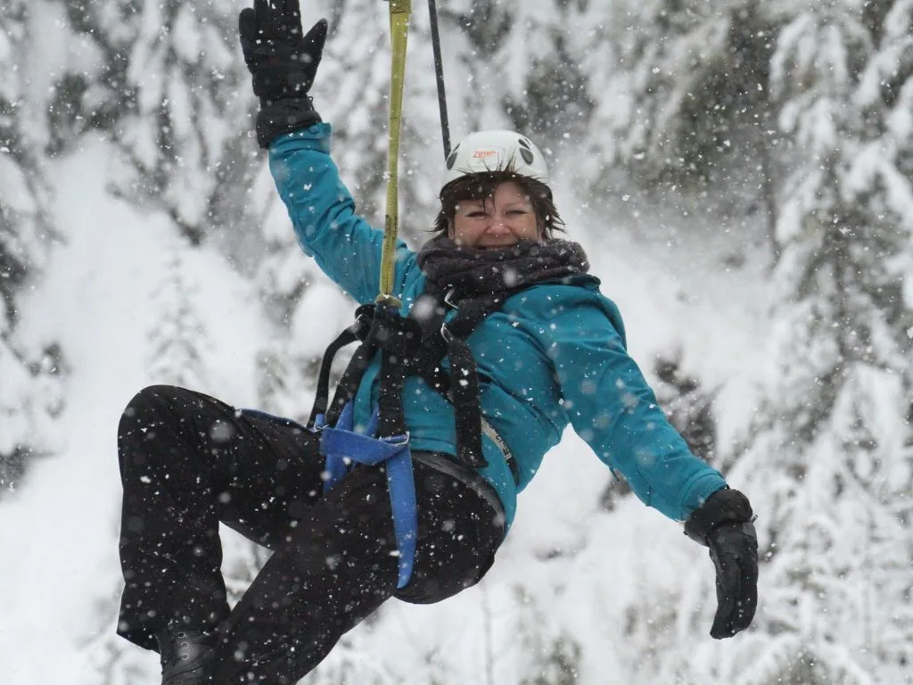 a woman wearing a blue coat is ziplining in whistler in the winter while snow is falling