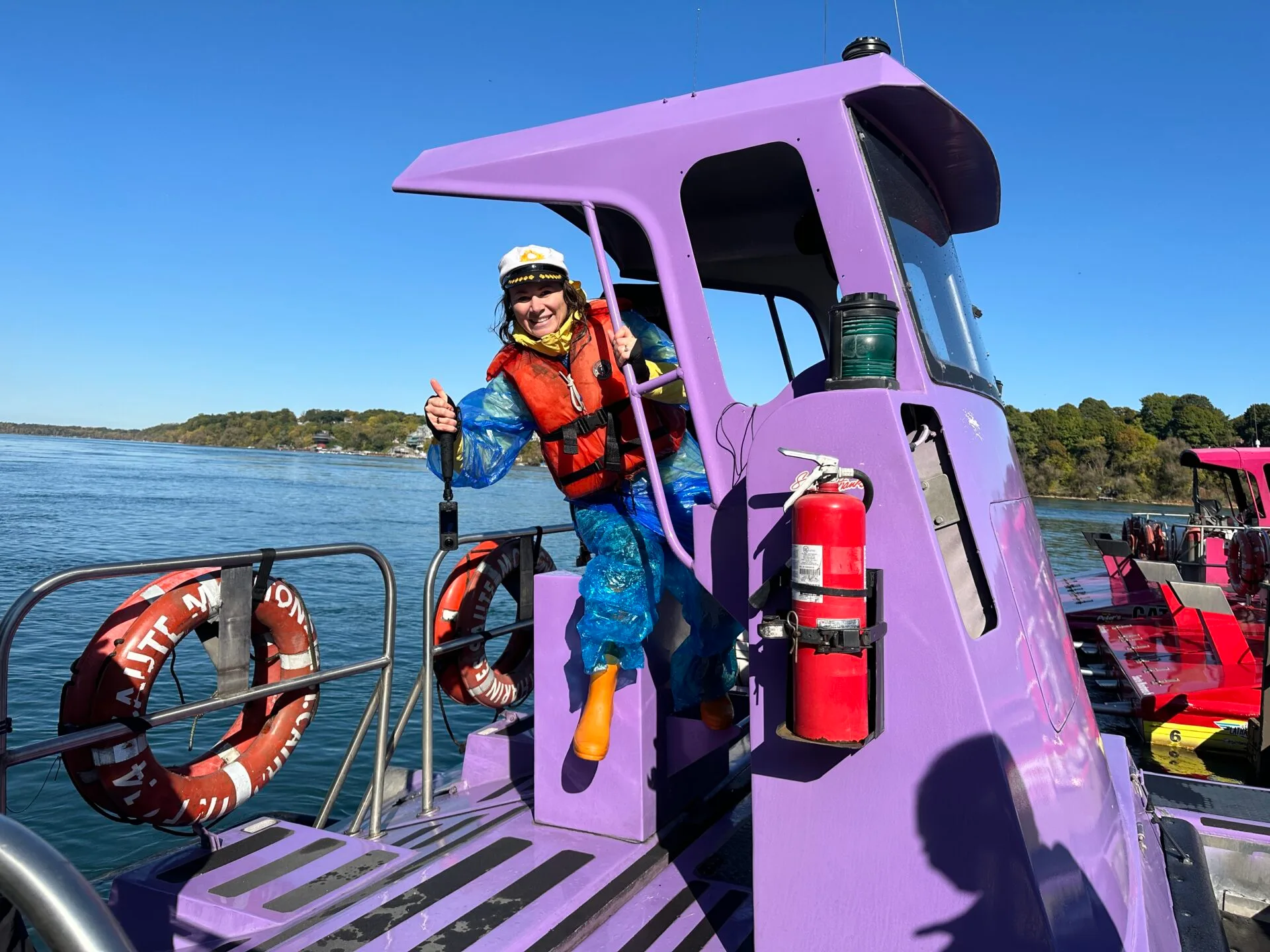 a woman wearing a captains hat, lifejacket, rain gear and orange boots sits in the captains seat of a purple whirlpool jet boat in Niagara Falls in October