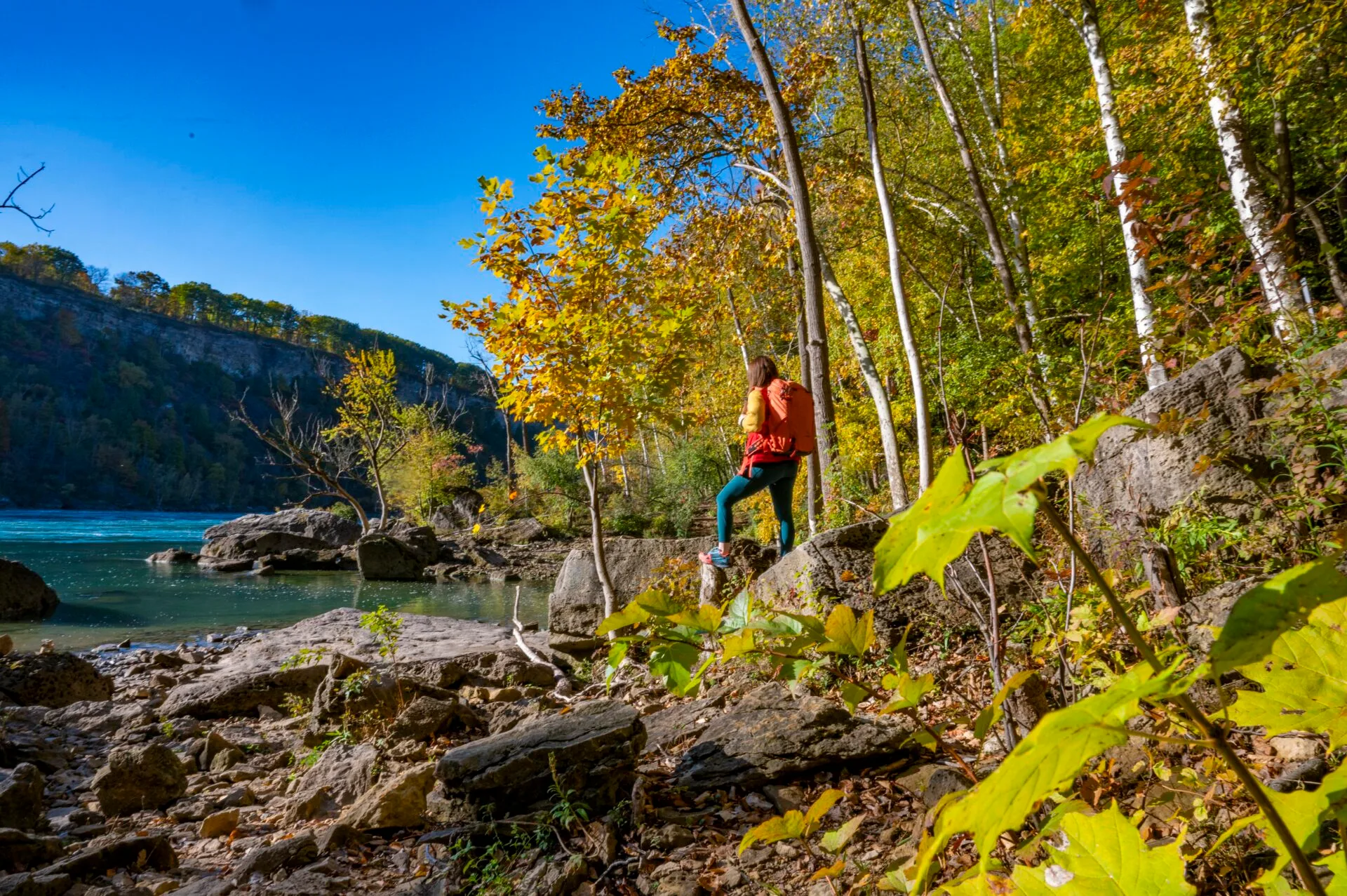 woman stands on boulder, wearing an orange hiking backpack, looking out at the niagara river while hiking in the niagara glen