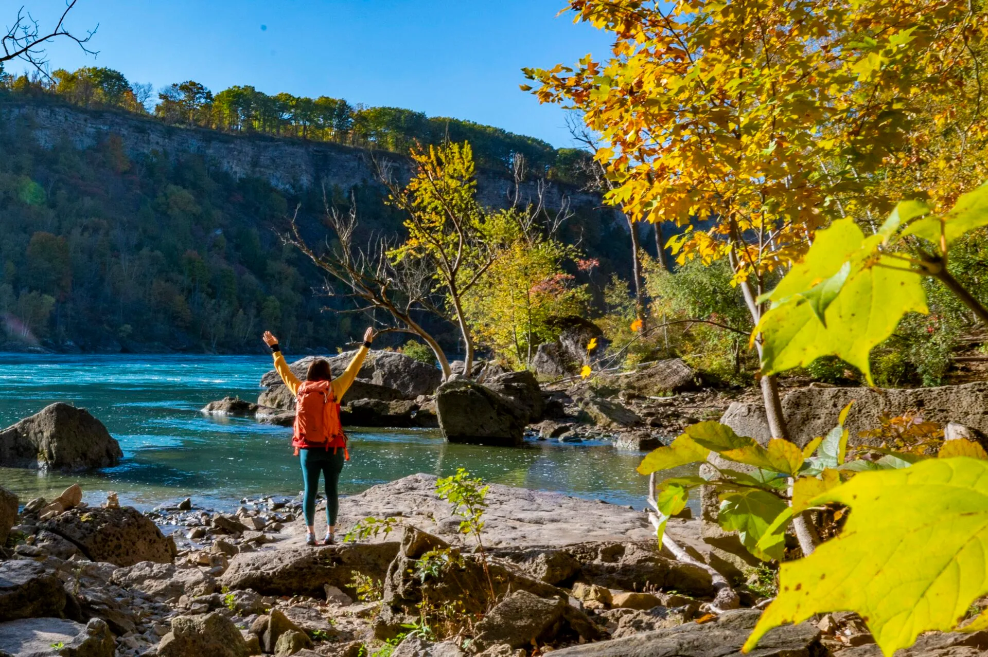 a woman stands on a large rock at the edge of the niagara river while hiking in the niagara glen