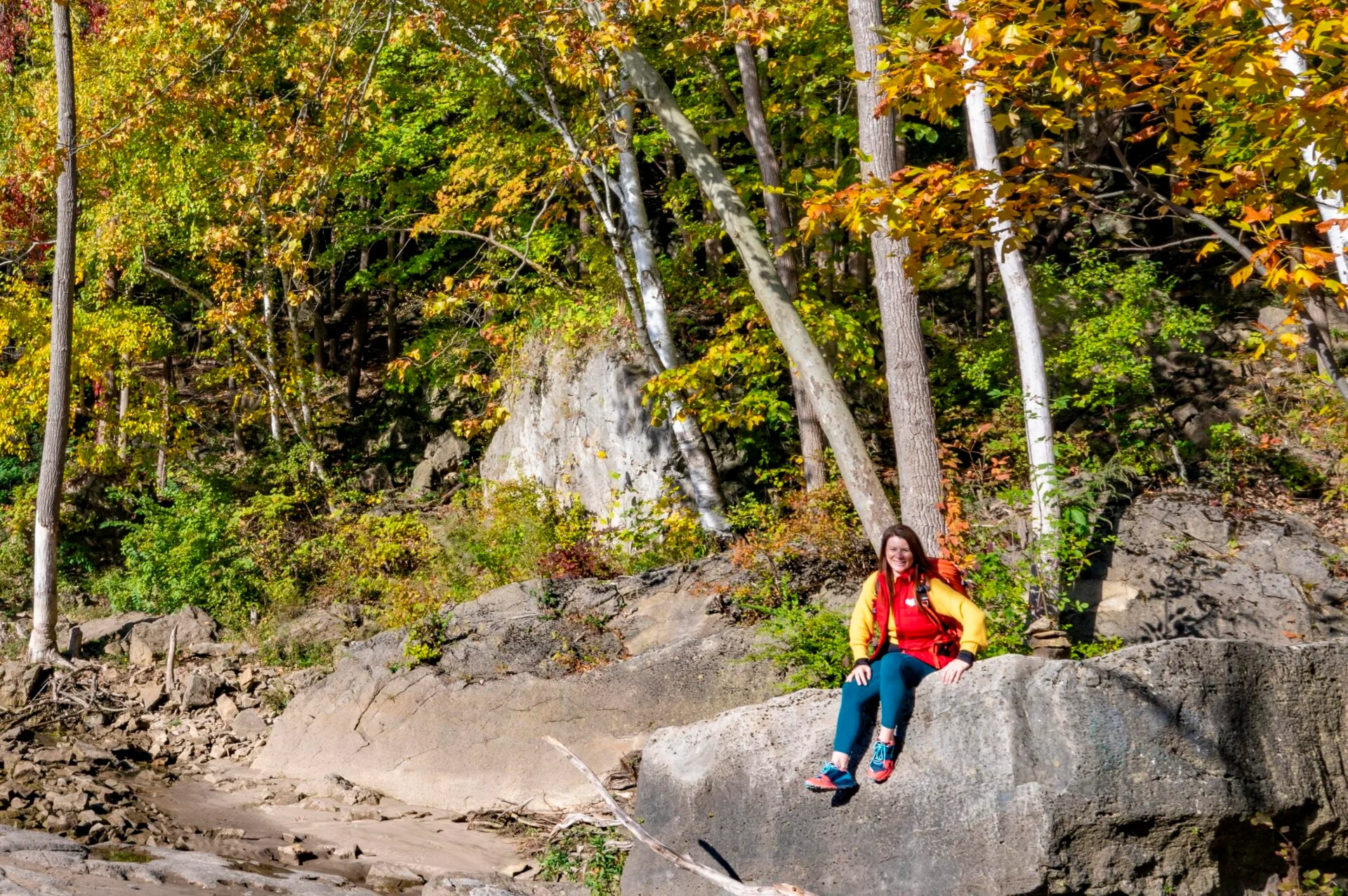 woman wearing a red vest and yellow sweater sits on a large rock and takes a break while hiking in the niagara glen