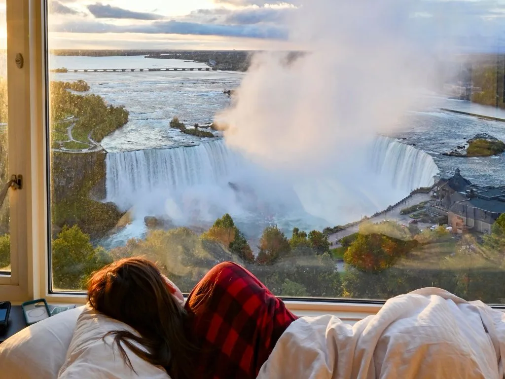 woman lying in a hotel room bed looks out the window at niagara falls