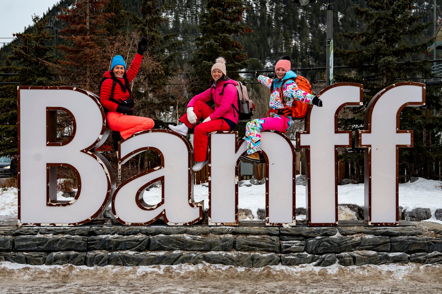 3 women wearing outdoor winter wear sitting on the large banff sign at the entrance of town