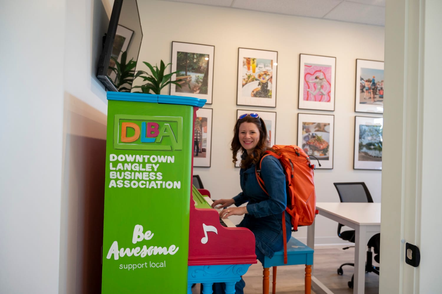 a woman with long brown hair and wearing an orange backpack its at a piano in the downtown langley business association's office