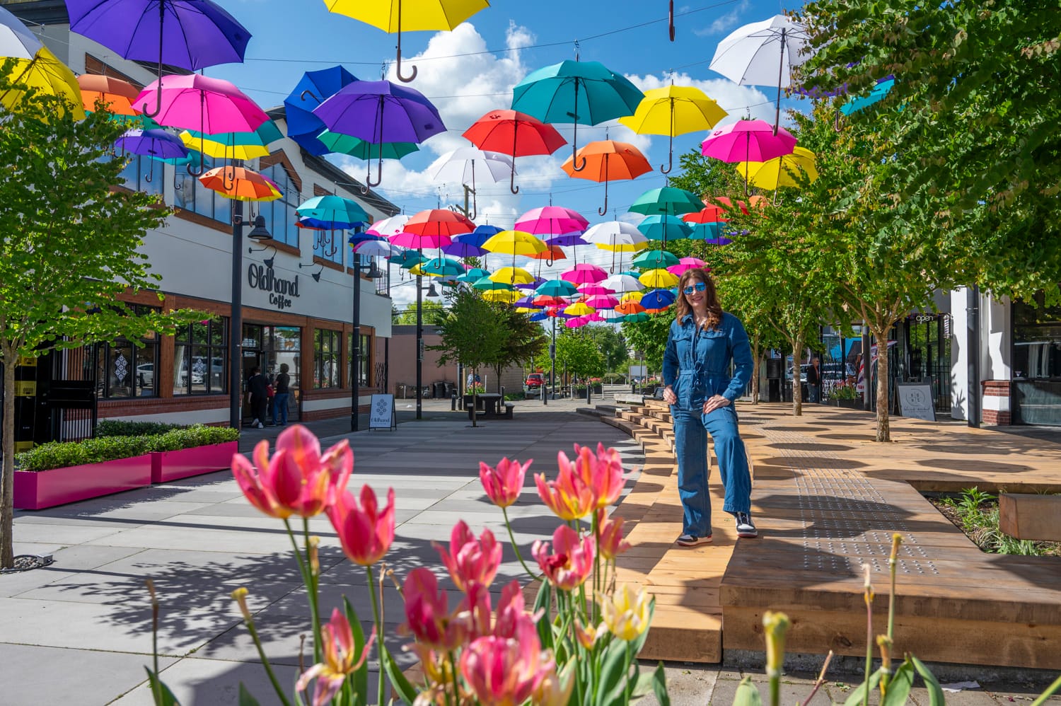a woman stands in mcburney plaza, it is a bright sunny day with blue skies and white clouds, colourful umbrellas are hanging from wires above the plaza