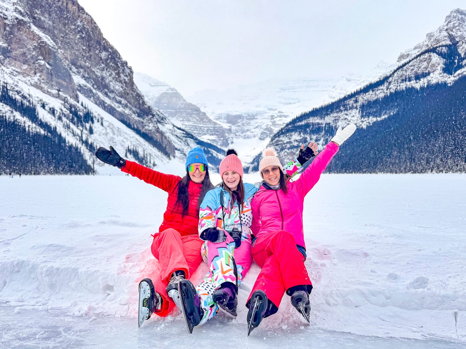 3 women wearing ice skates and sitting on the snow and ice on lake louise in alberta