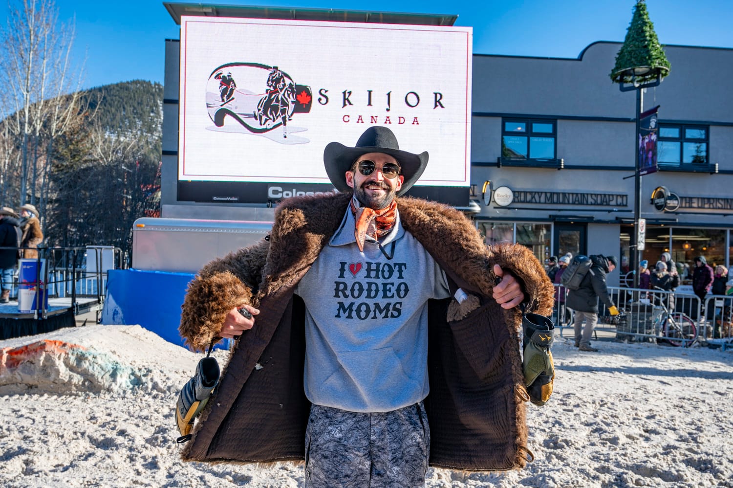 a man wearing a cowboy hat and furry brown winter coat holds it open to reveal his sweatshirt underneath says I heart rodeo moms