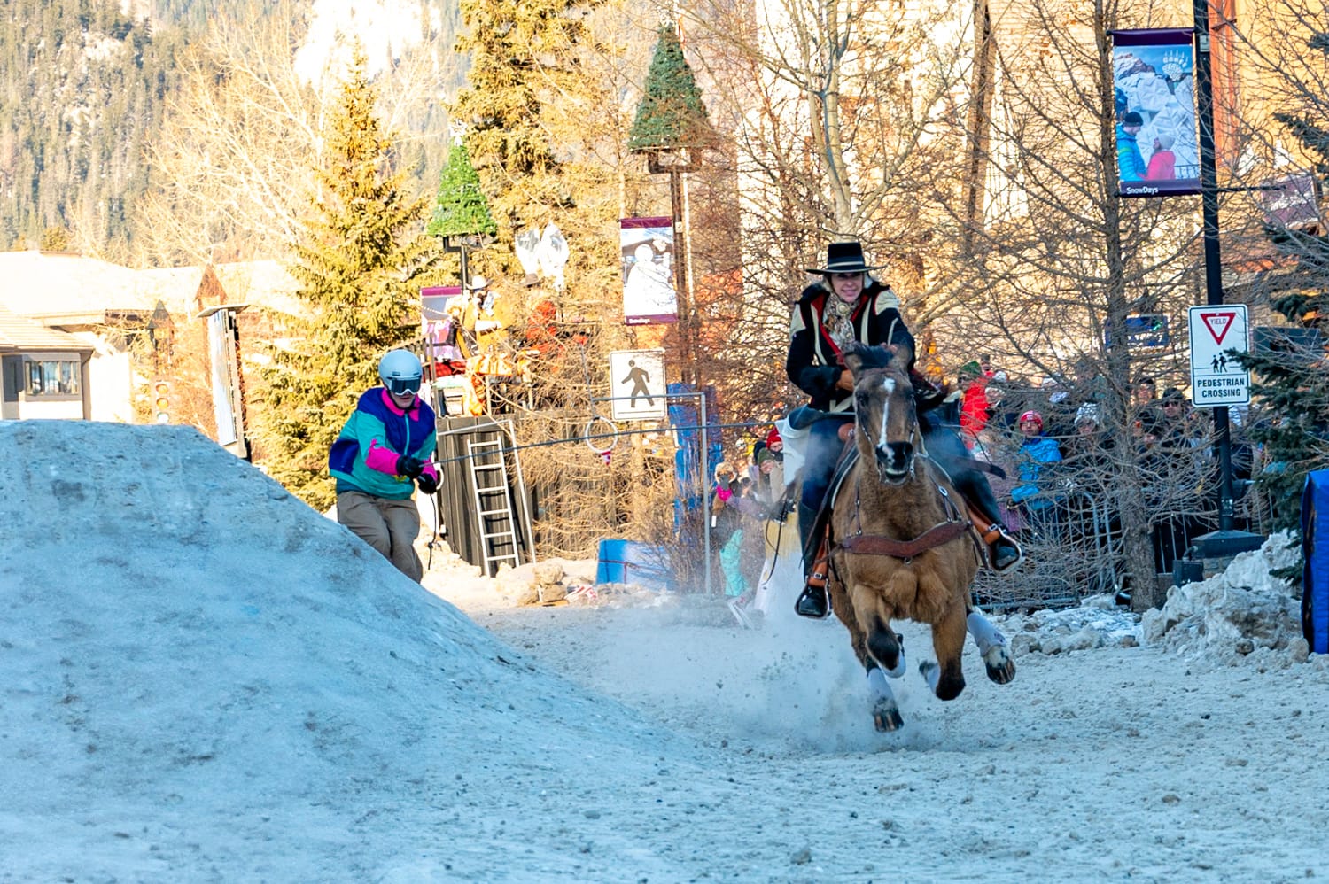 a woman rides a brown horse, it is running towards the camera and is pulling a skier behind it on a rope