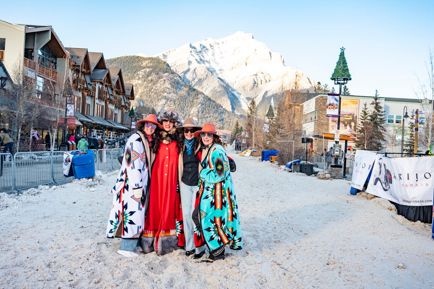 four women, wearing western clothing, stand on the snow in the middle of a banff street during a skijoring event
