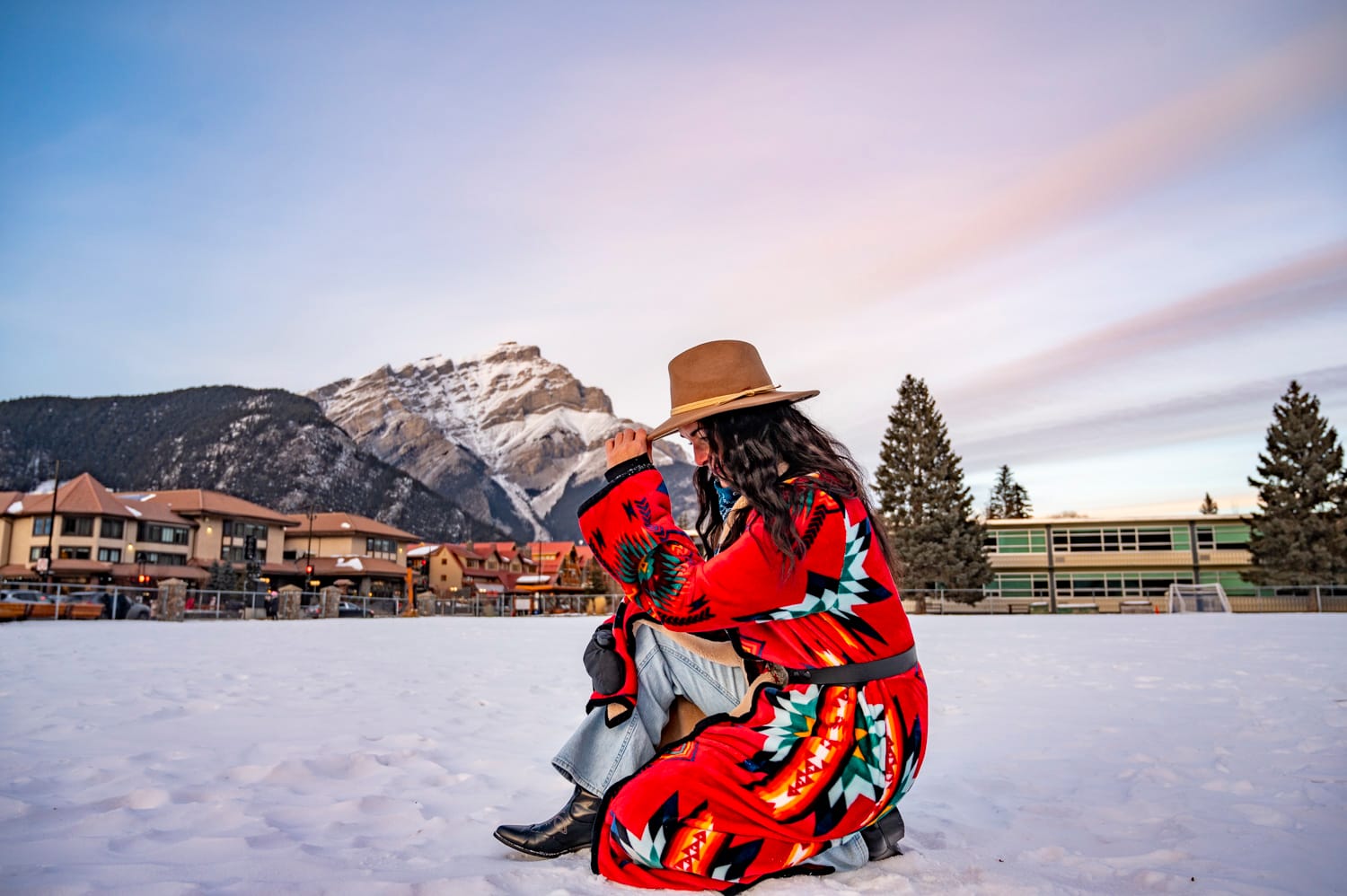 a woman wearing a red western coat, brown cowboy hat, jeans and boots, crouches down in the snow, the town and mountains of banff stand in the background