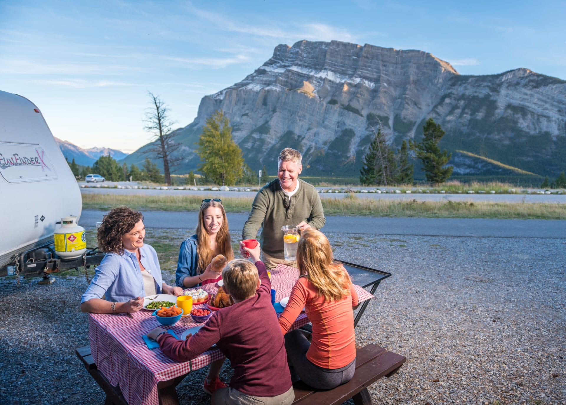 a family sits at a picnic table beside their RV trailer at tunnel mountain village II campground, a large rocky mountain is behind them