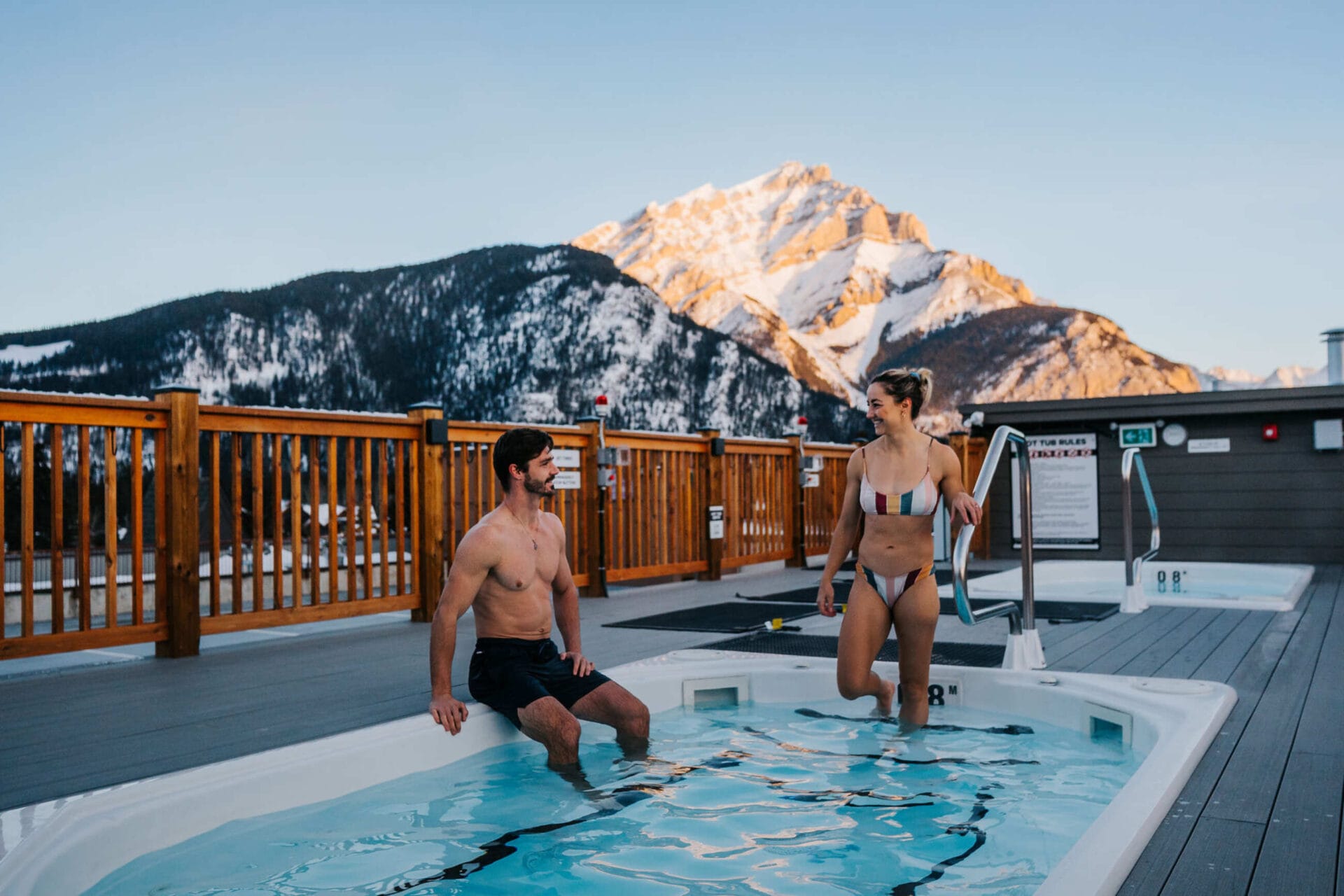 a couple is walking into a roof top hot tub at basecamp suites banff, a mountain in the background
