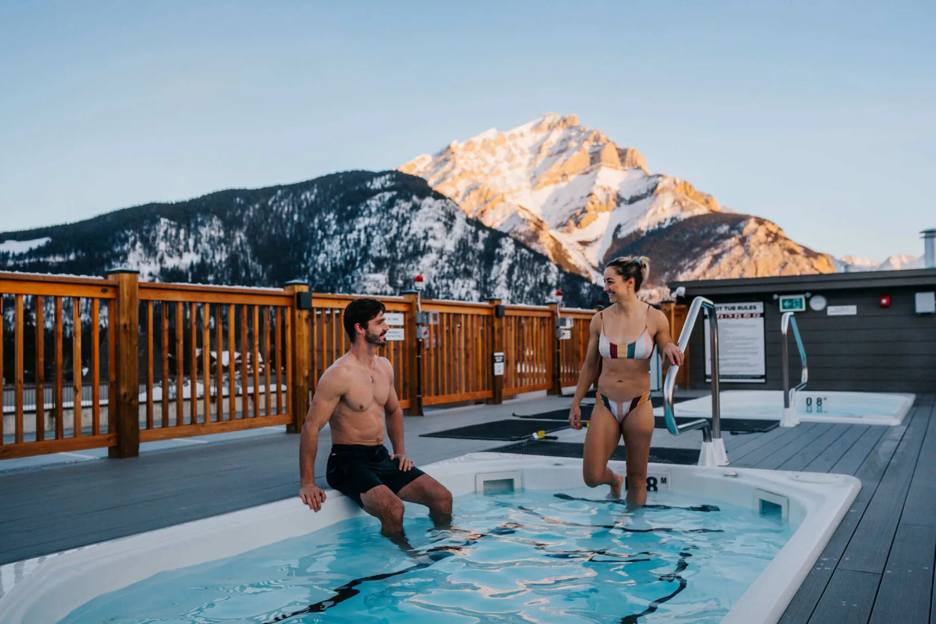 a couple is walking into a roof top hot tub at basecamp suites banff, a mountain in the background