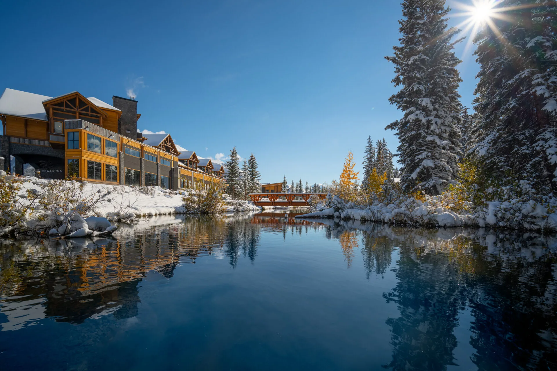 the malcolm hotel sits behind a pond and forest, a wooden bridge is off in the distance, the ground is covered with snow